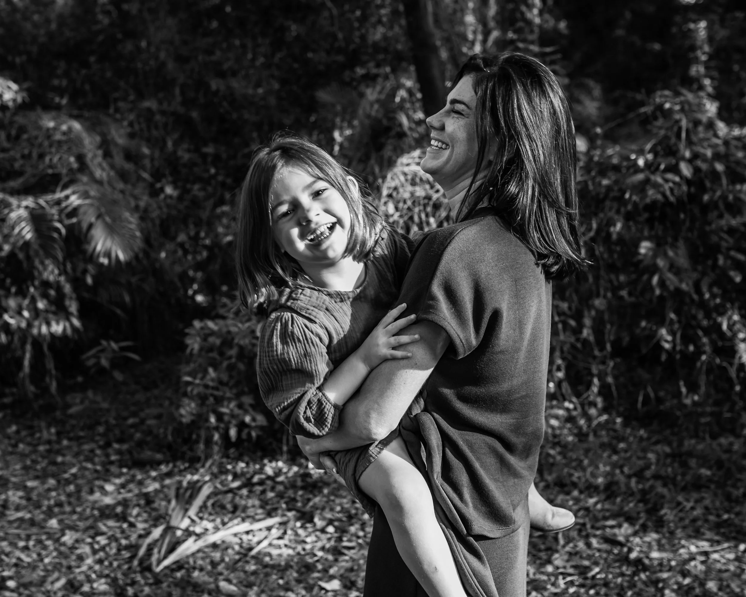 A woman lifting a young girl in a park, both smiling and enjoying the moment. The woman is holding the girl close, and they are surrounded by trees and foliage.