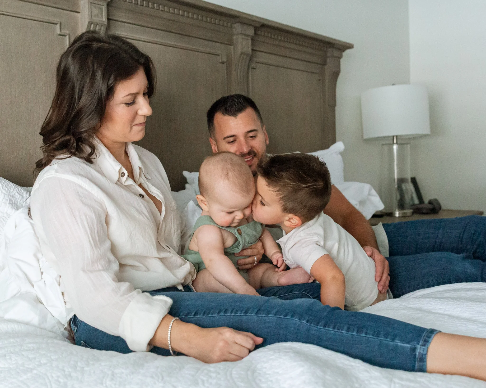 A family of five sitting on a bed, with the mother and children kissing while dad smiles in the background. The mother has dark hair, and the three children have light hair. The bed has a white quilt, with a beige headboard and a white wall with a la