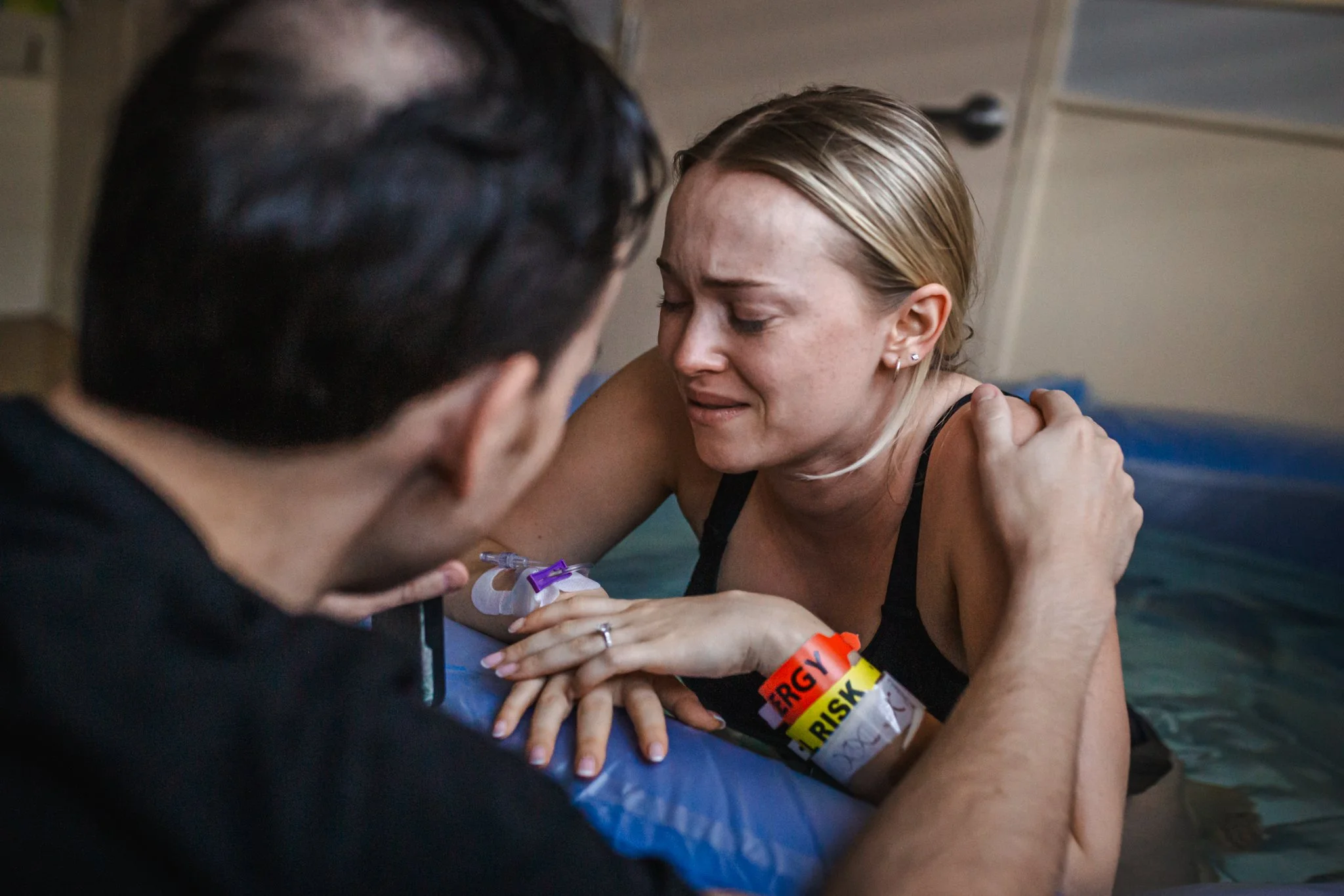 A woman with blonde hair and light skin, visibly emotional and crying, is being comforted by a person with dark hair. The woman has an intravenous (IV) line in her hand, indicating she is in a medical setting, likely giving birth or recovering from c