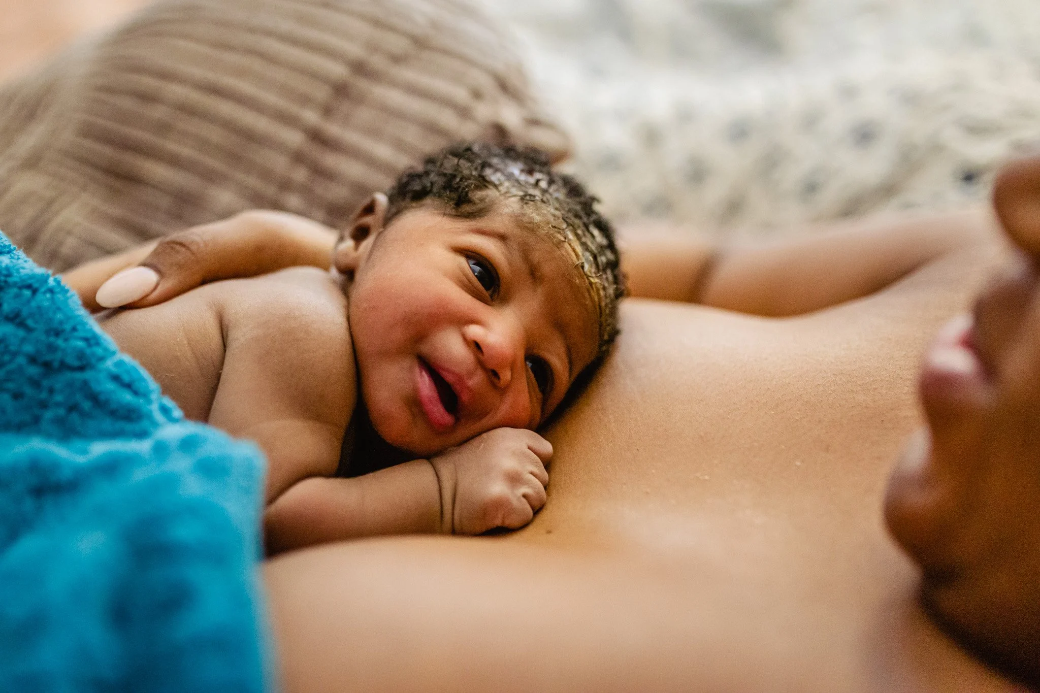 Close-up of a smiling baby lying on a person's chest with a blanket nearby.