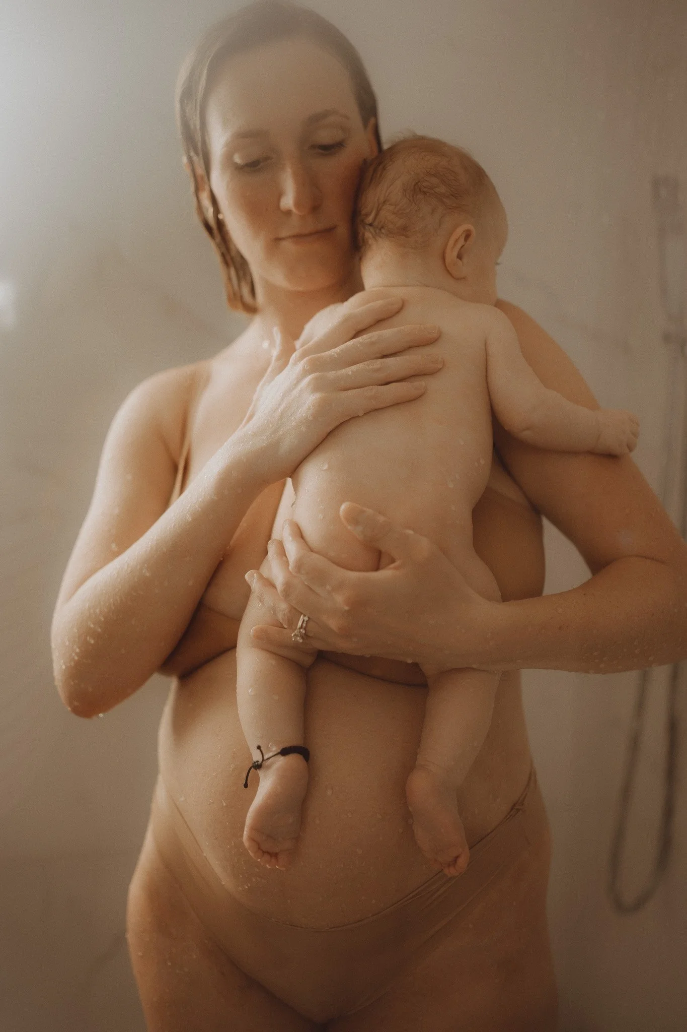 A woman with short brown hair is holding a naked baby against her chest, with the baby's head resting on her shoulder. The woman appears to be in a shower or bath, with water and soap on her skin. The background is neutral and out of focus.