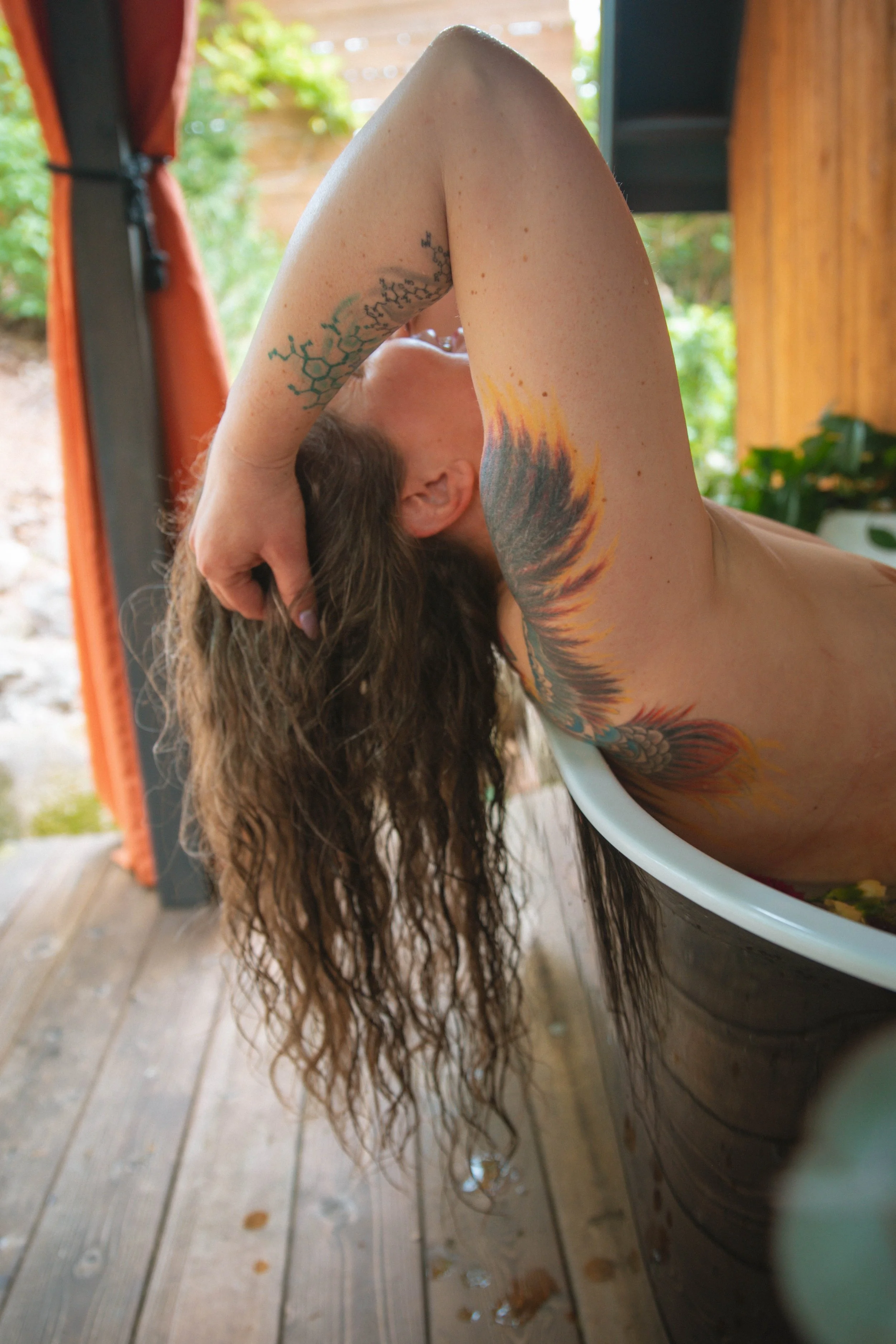 Person lying in a bathtub with long, curly hair hanging out, with colorful tattoos on arm and shoulder, on a wooden outdoor deck with greenery visible outside.