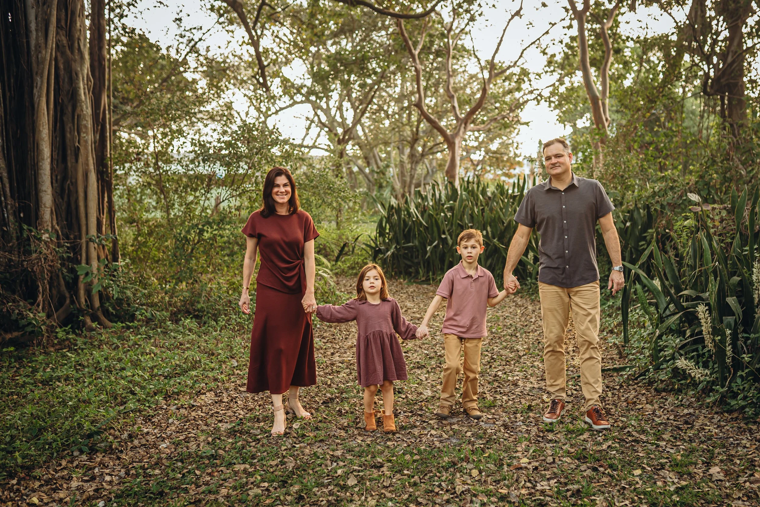 A family of five walking through a wooded park. The mother wears a maroon dress, the father wears a gray shirt and khaki pants, and three children wear earth-toned clothes. They are holding hands and smiling as they walk along a leaf-covered path.