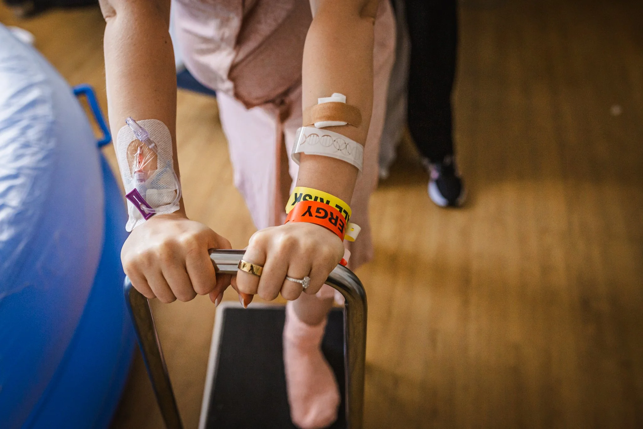 Close-up of a person's arms and hands, with one hand gripping a metal railing. The person has bandages, medical tubing, and several wristbands on their wrists, indicating they are a hospital patient.
