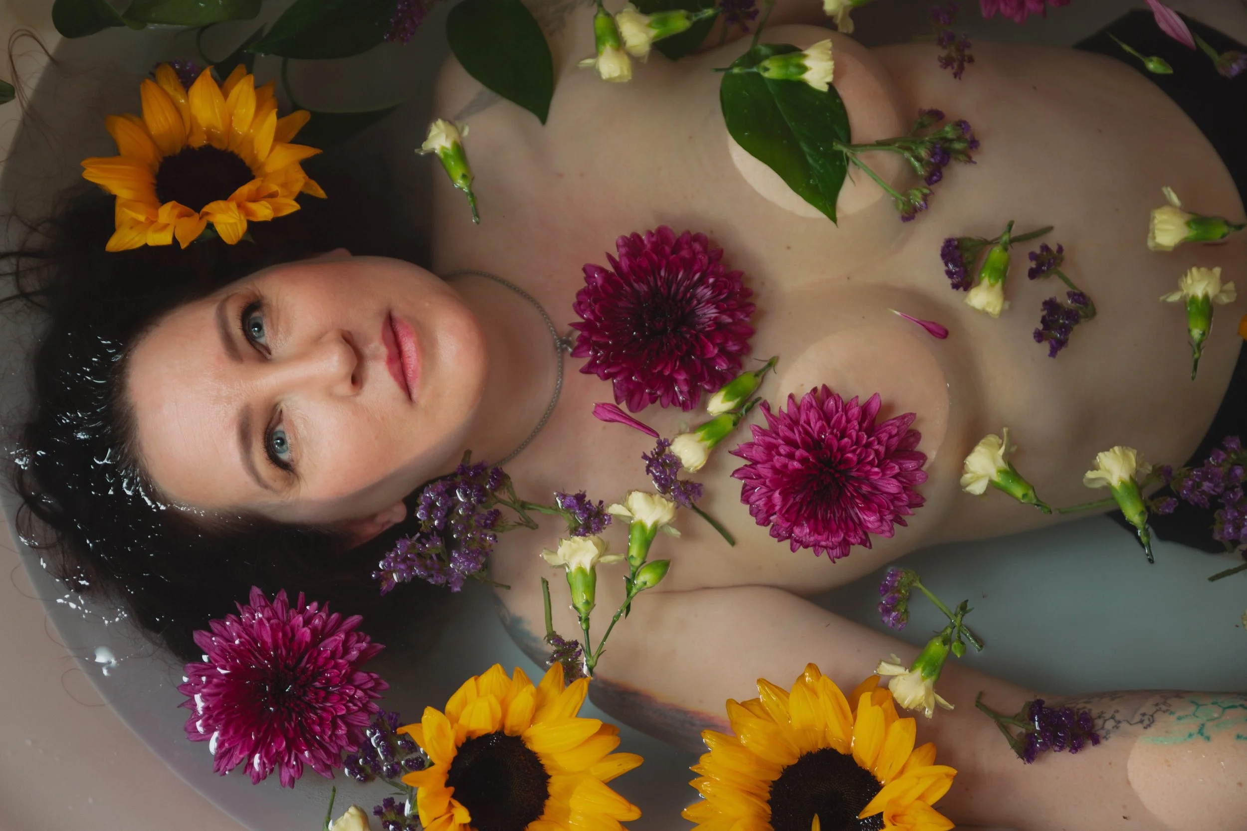 A woman with dark hair and blue eyes lying in a bathtub decorated with various colorful flowers, including sunflowers, pink chrysanthemums, and small purple and white flowers, with a relaxed facial expression.