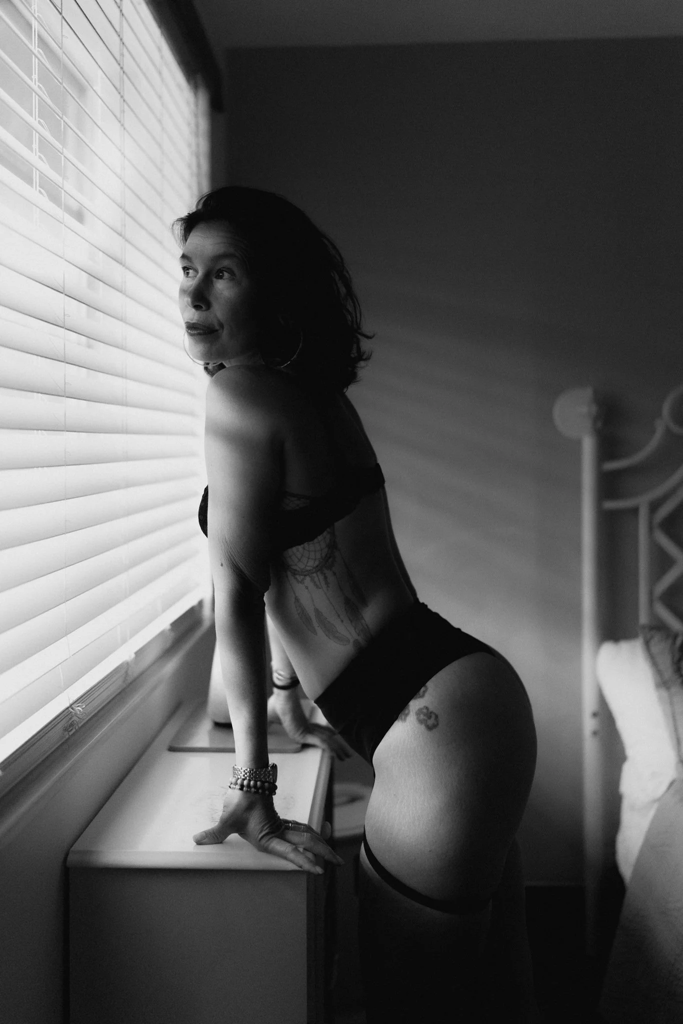 Black and white photo of a woman leaning on a windowsill, looking out the window with blinds, wearing lingerie, tattoos, and jewelry.