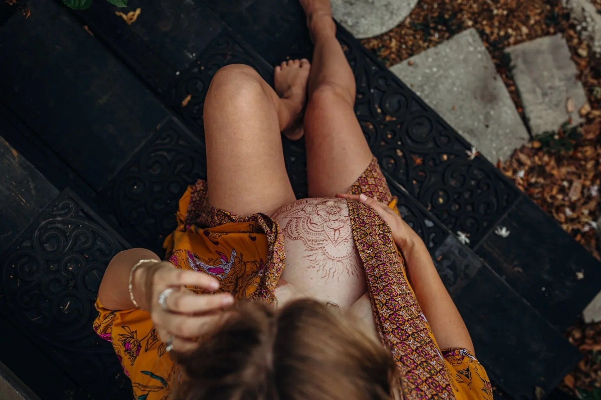 A person lying on their back, wearing an orange dress with a floral pattern, with a tattoo on their abdomen visible. Their left hand is resting on their abdomen, and a person near their head is reaching towards them. There are stone and concrete steps nearby, and fallen leaves on the ground surrounding them.