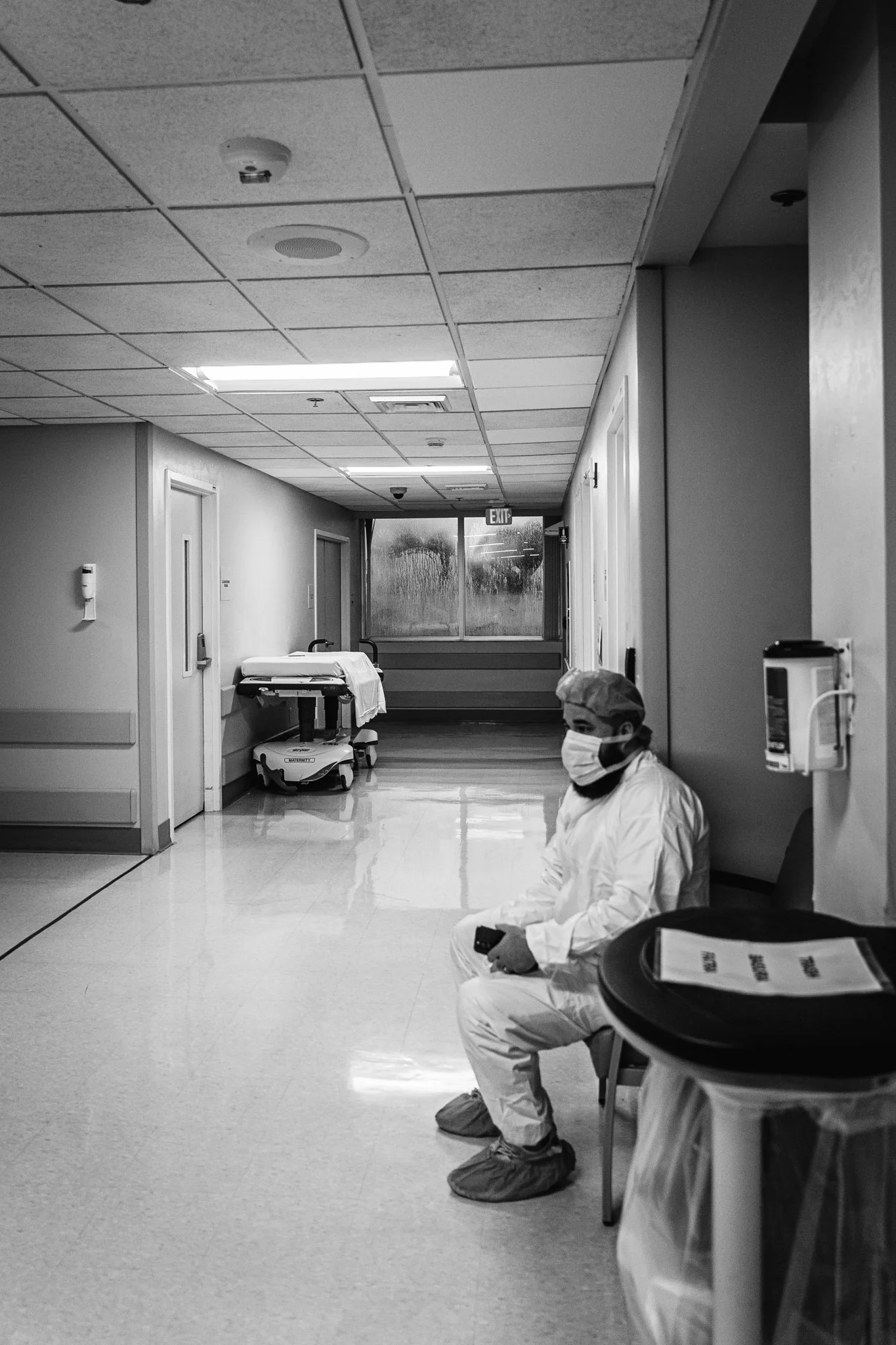 A person wearing white medical scrubs, a mask, and shoe covers sitting in a hospital corridor, holding a phone. The corridor has closed doors, hospital equipment, and an exit sign visible at the end.