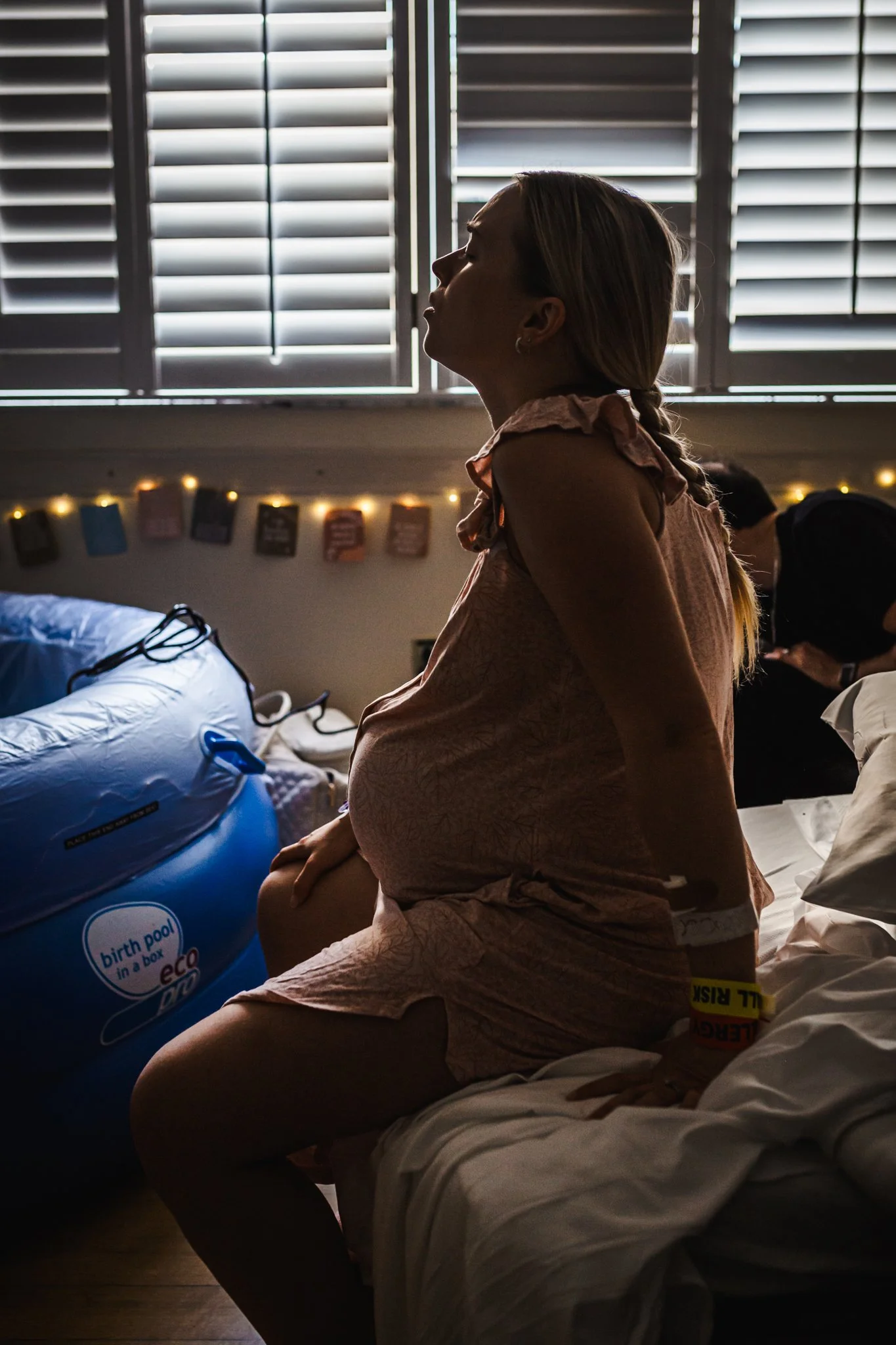 A pregnant woman sitting on a hospital bed, wearing a hospital gown, with her hands clasped on her lap and a yellow wristband, in a dimly lit room with blinds and string lights in the background.