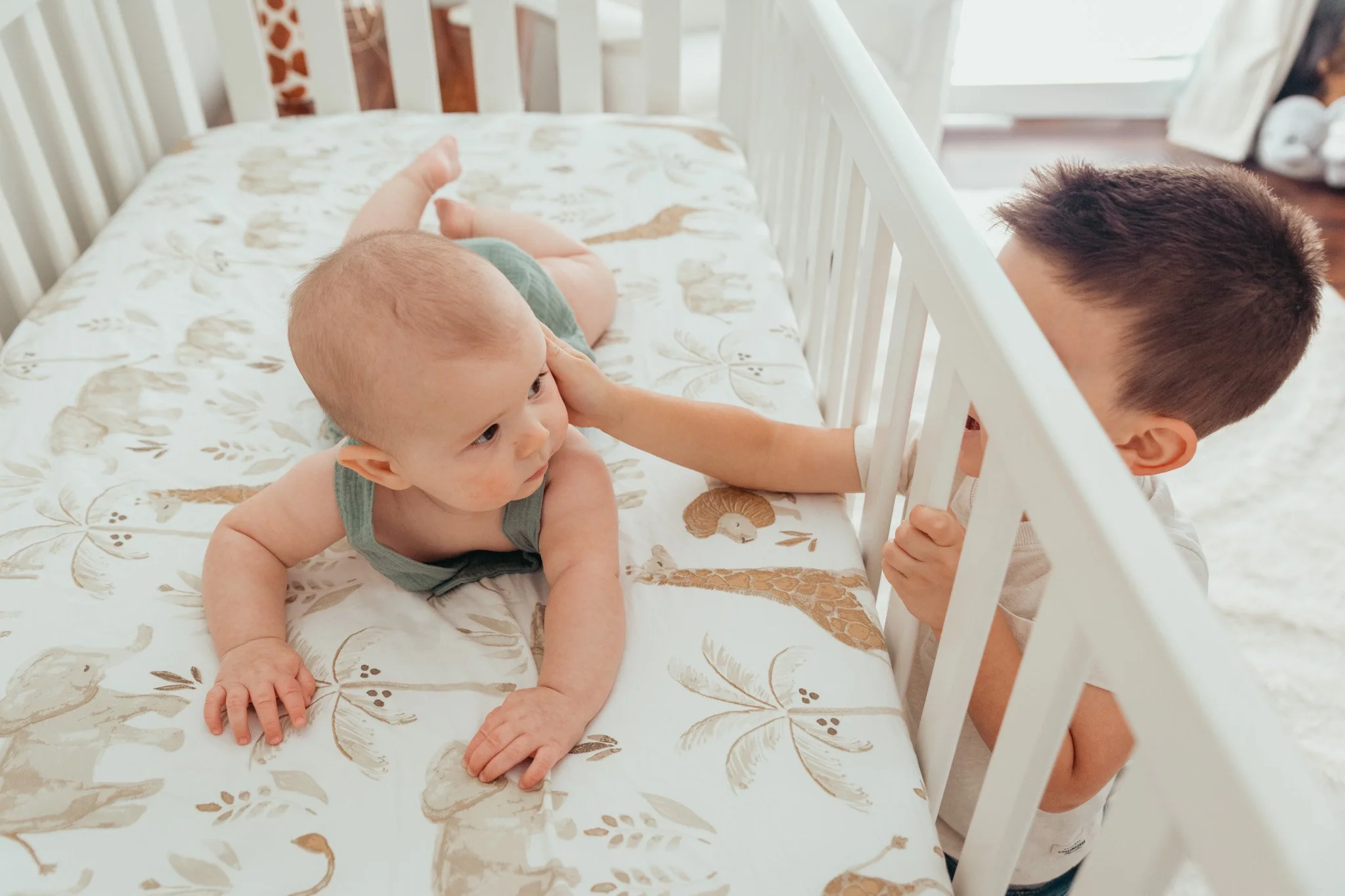 A young boy with short dark hair leaning over a white crib, looking at a small baby lying on a bed with a floral patterned sheet, touching the baby's face gently.