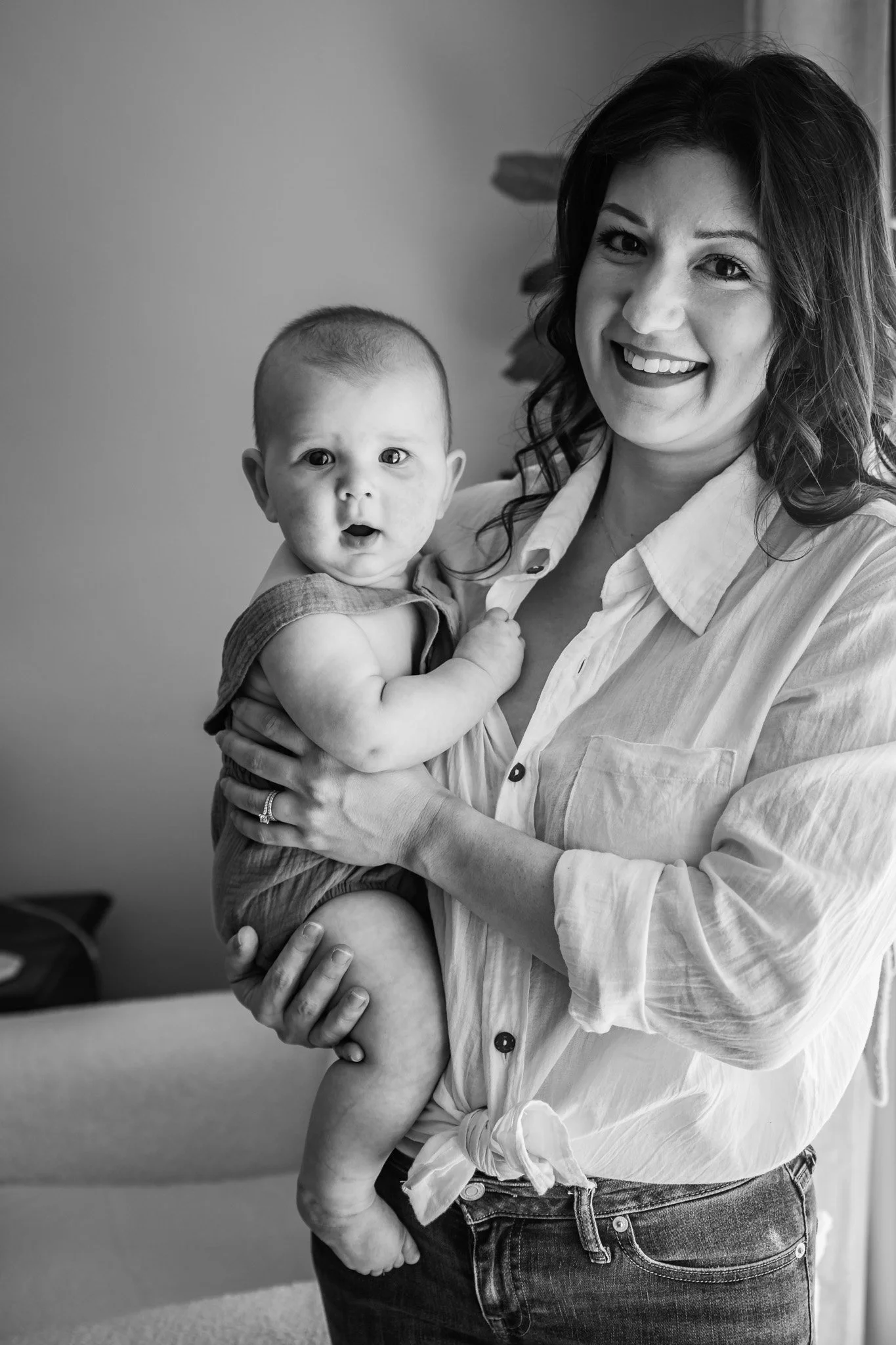 A woman with dark hair smiling and holding a baby in her arms in an indoor setting.