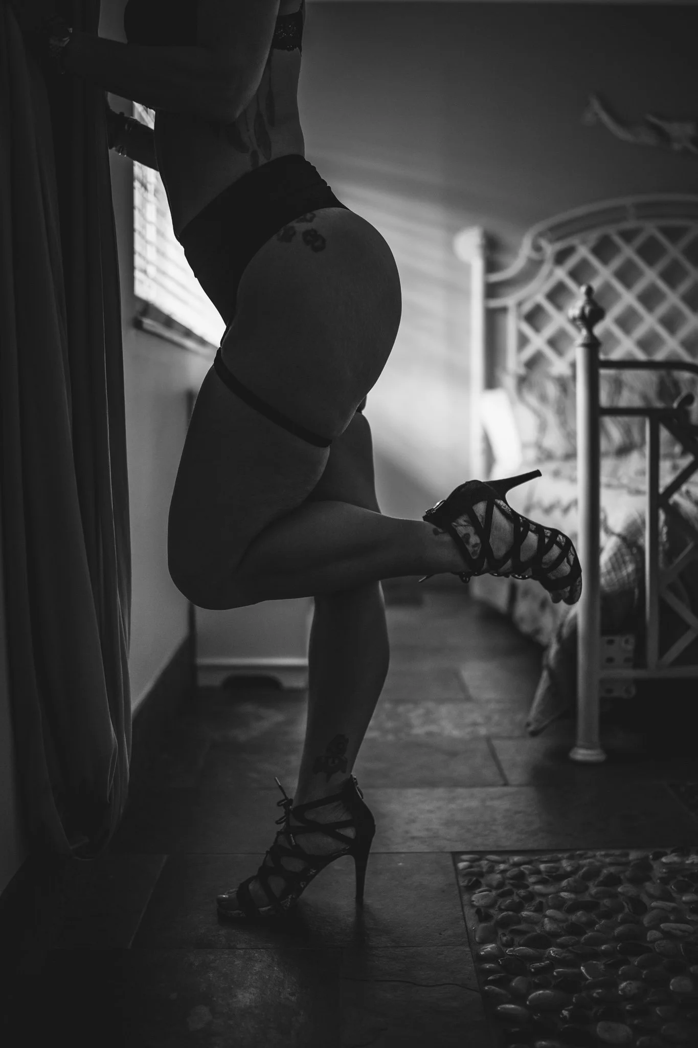 Black and white photo of a woman standing in profile, wearing high heels, patterned stockings, and lingerie, leaning against a piece of furniture in a dimly lit room.