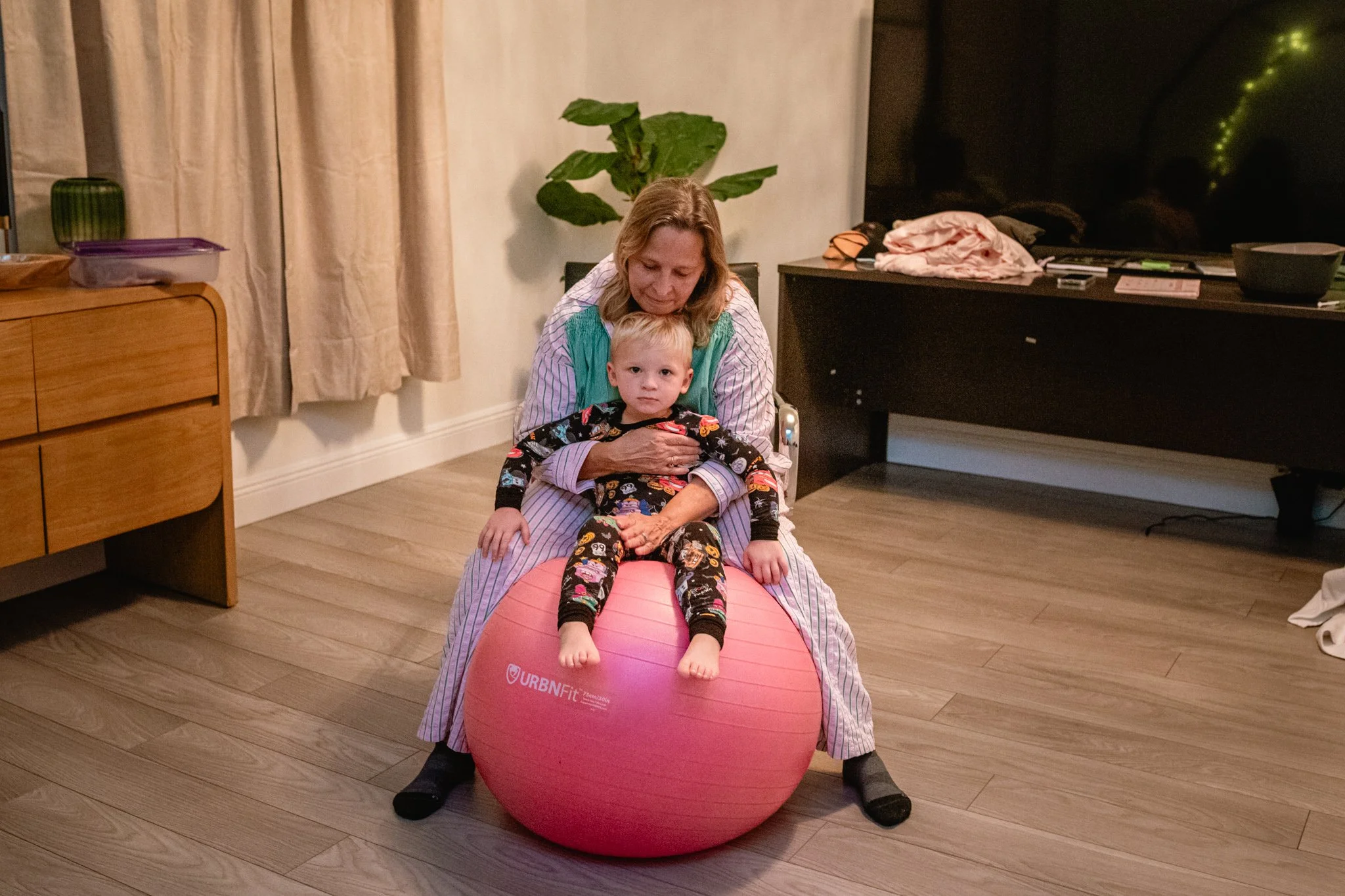 A woman and a young boy sitting on an exercise ball in a living room. The woman is behind the boy, with one hand on his stomach, helping him balance on the pink exercise ball. The boy is wearing pajamas with a colorful pattern, and the woman has long