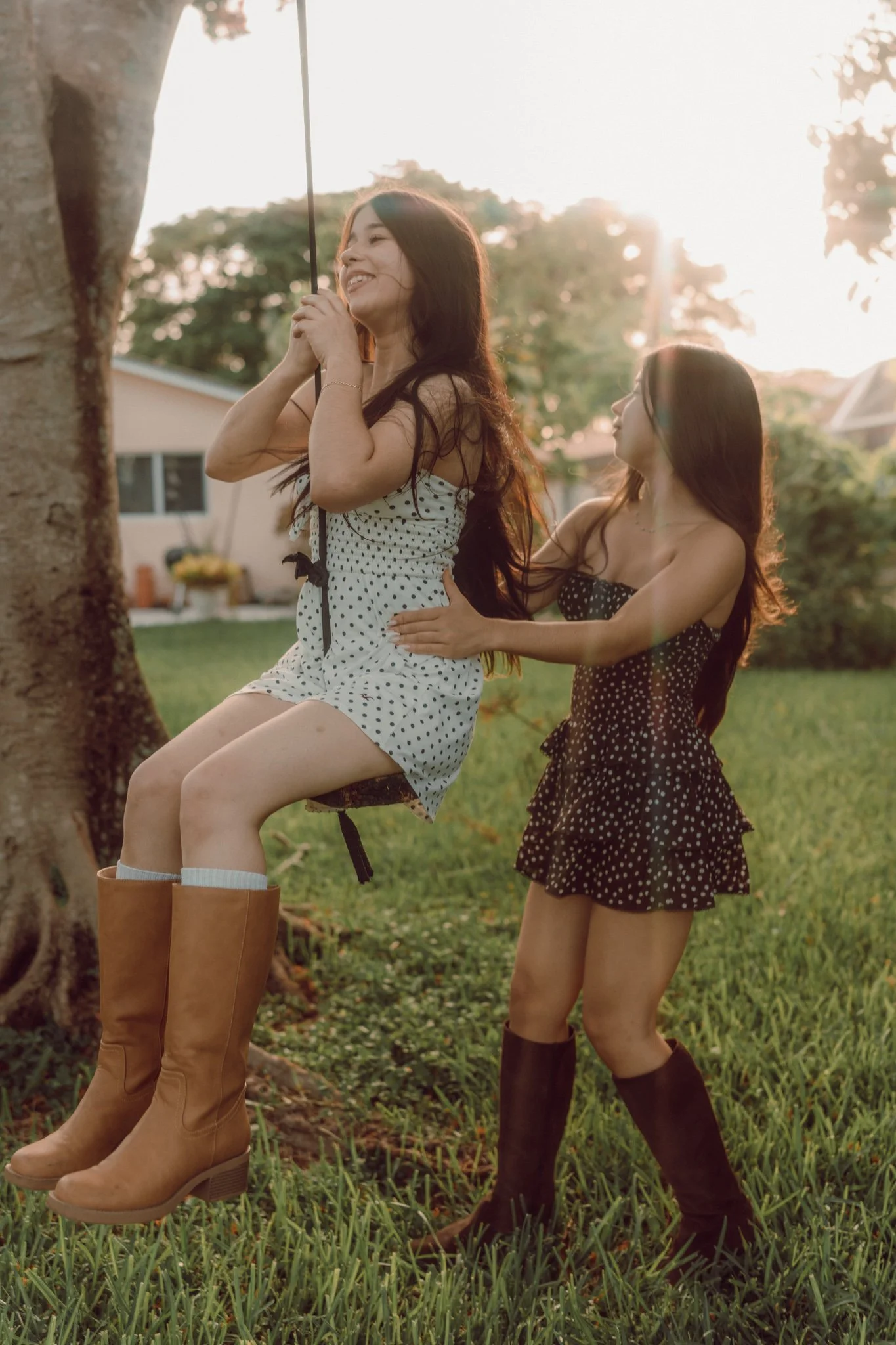 Two women outdoors near a tree, one sitting on a swing and smiling, the other standing beside her, with sunlight filtering through trees in the background.