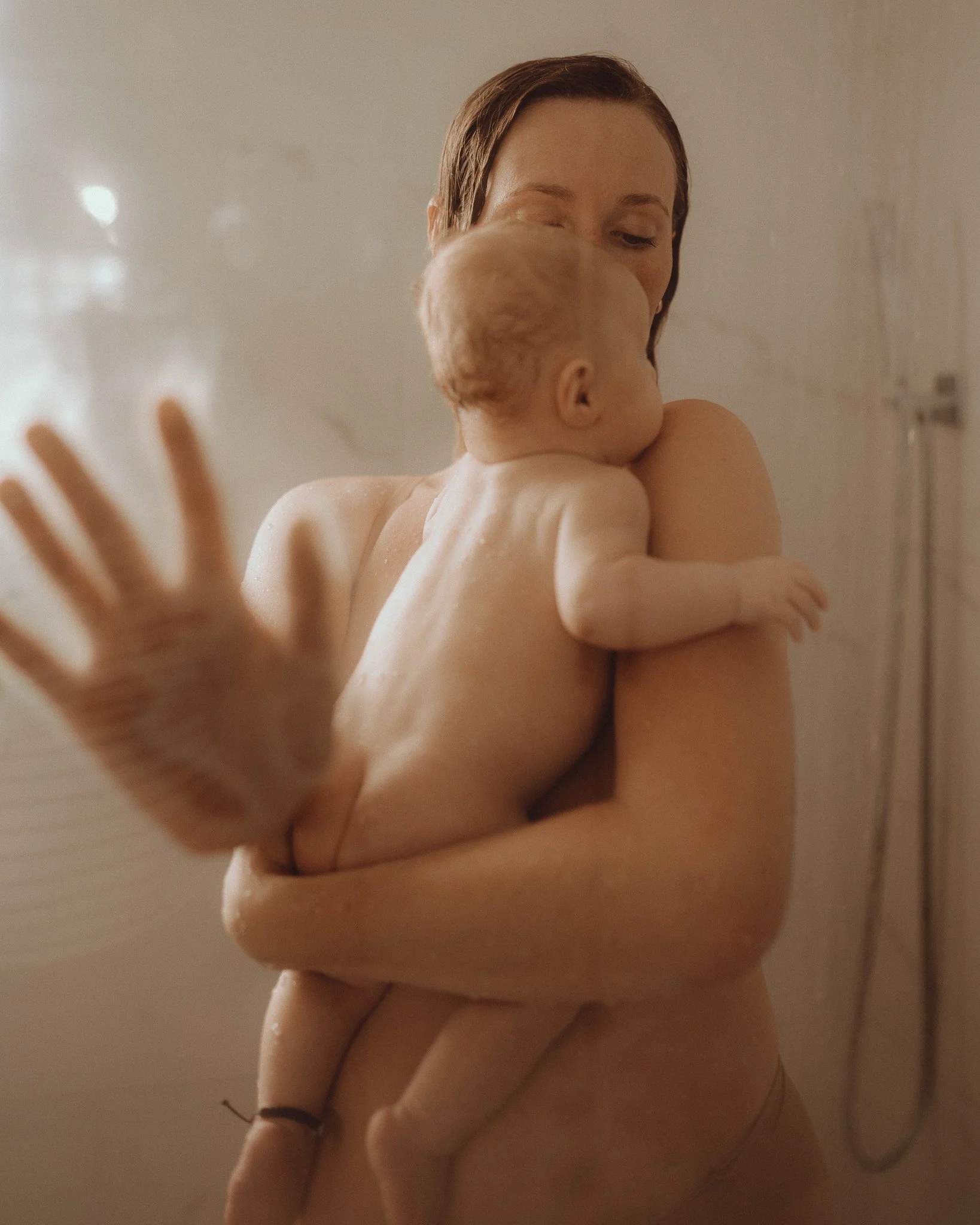 Close-up of person with wet hair holding a baby in a shower or bath, focusing on the back of the person's head and the baby's shoulder and head.