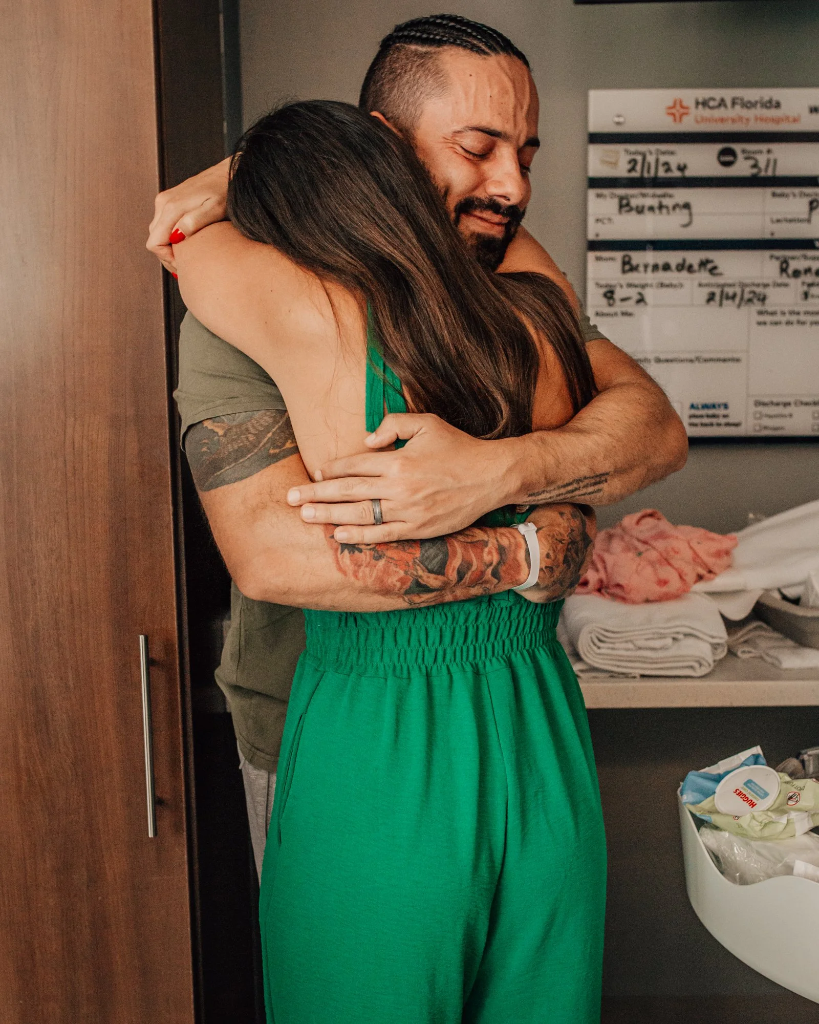 Two people hugging in a hospital room, with hospital supplies and a medical chart on the wall behind them.