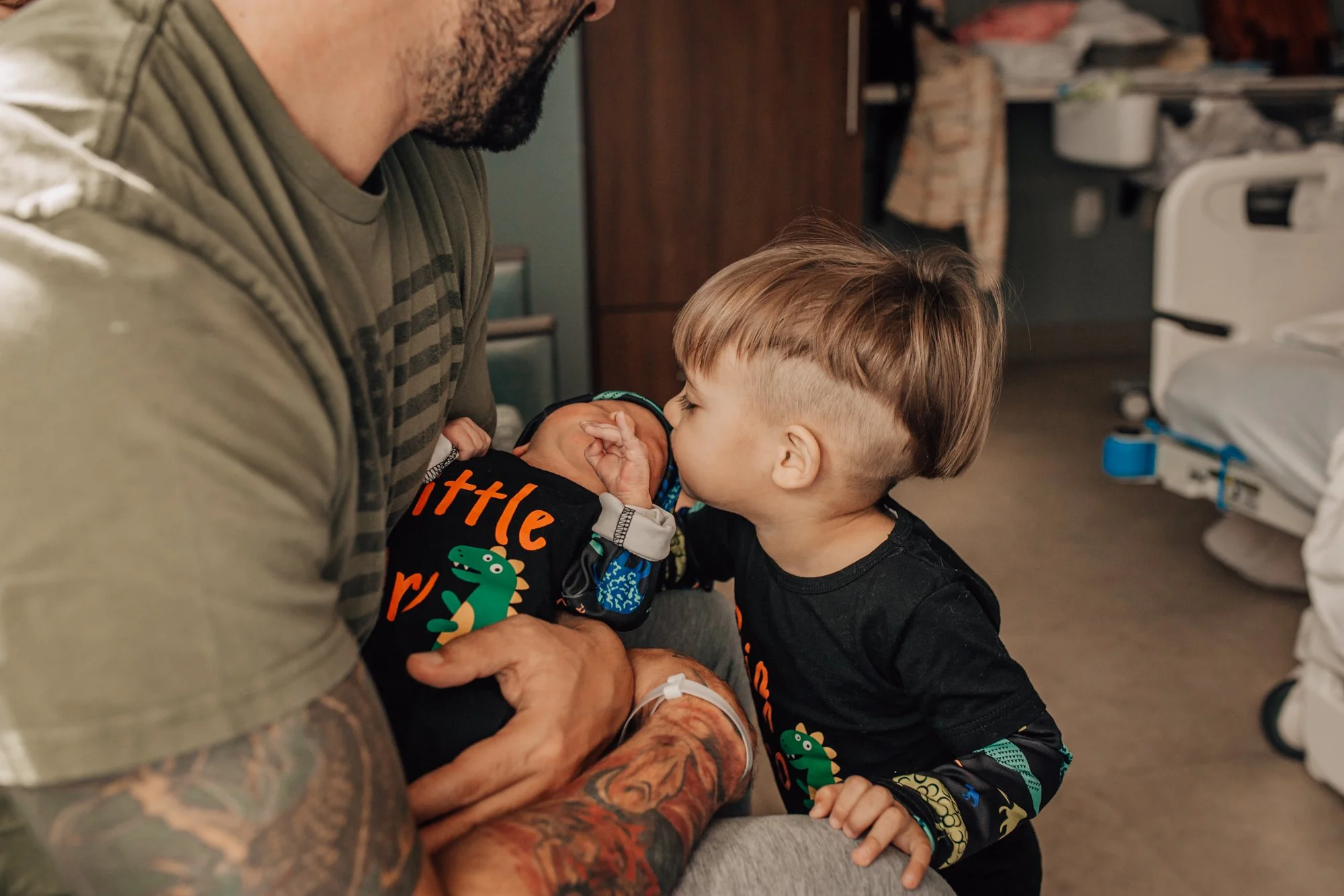 A young boy and an older man, both with dark hair, are close together in a hospital room. The boy is touching noses with a newborn baby, possibly his sibling, who is wrapped in a blanket with colorful dinosaurs. The man has tattoos on his arms and is
