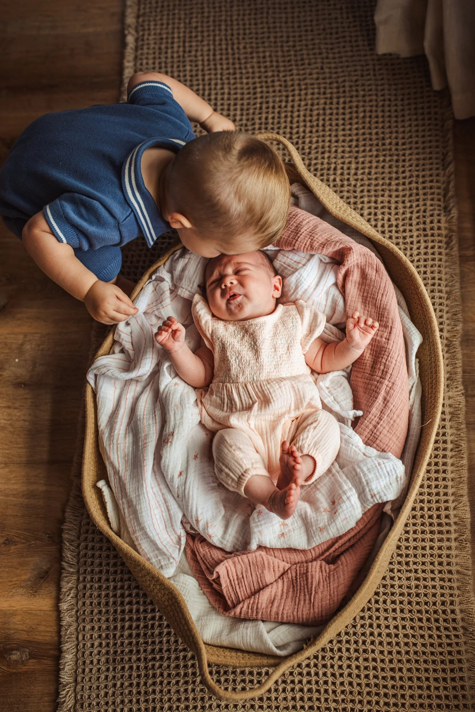A young boy with blond hair in a blue shirt leaning over a bassinet, kissing an infant baby who is lying down and making a funny face, with a woven brown blanket and a textured brown rug beneath them.