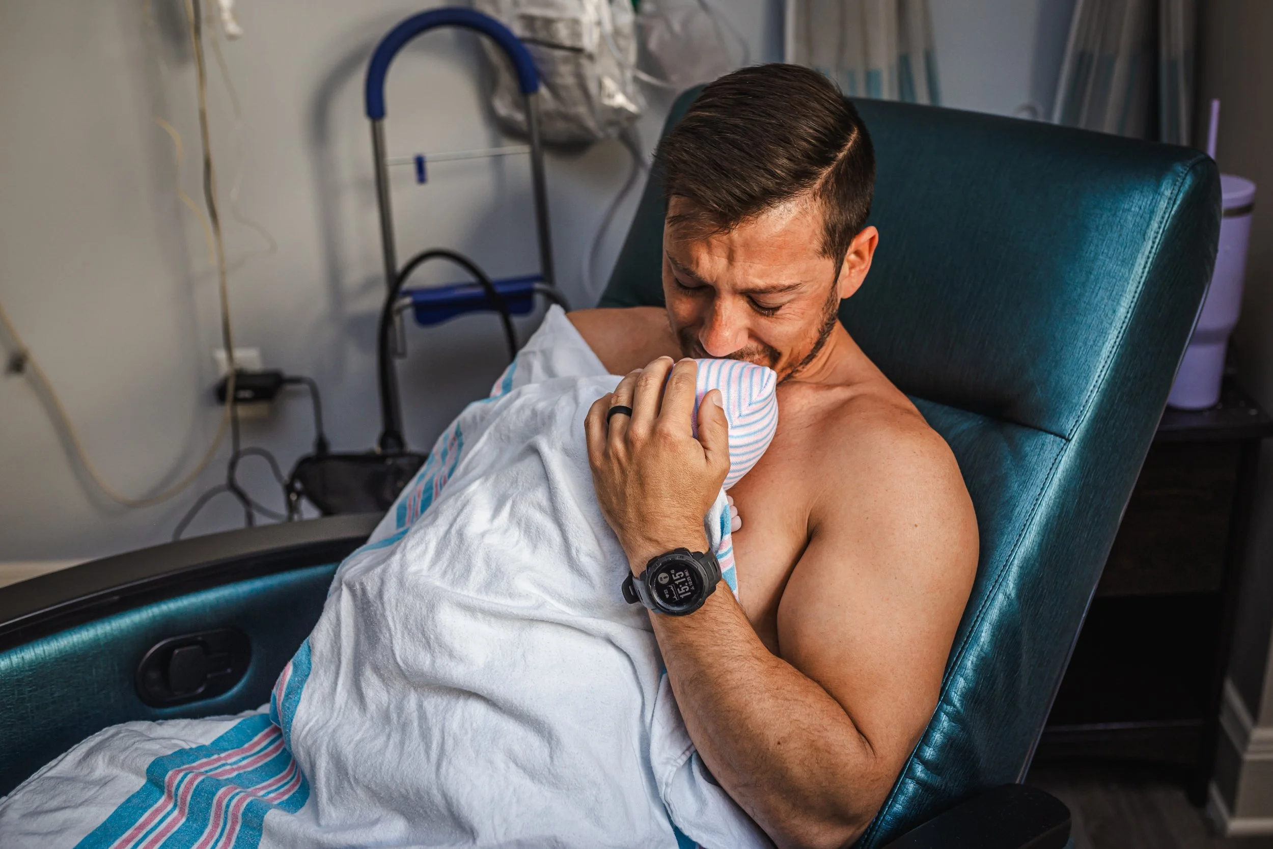 A man is lying in a medical chair in a healthcare facility, holding a newborn baby wrapped in a hospital blanket, gently touching the baby's face and smiling.