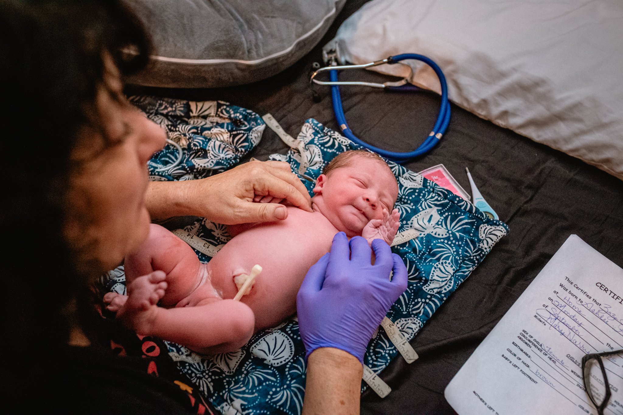 A newborn baby being born in a hospital. The baby is lying on a cloth with a blue and white pattern, with one hand under its chin and eyes closed. A woman, possibly the mother, is supporting the baby and a person wearing purple gloves is touching the