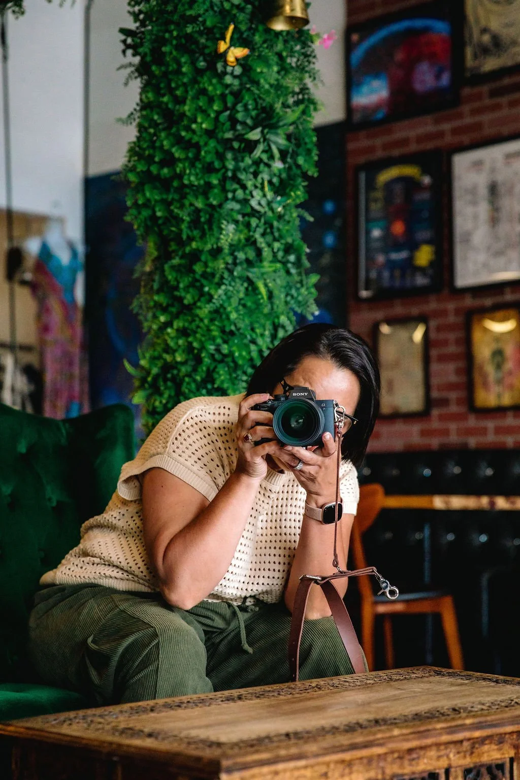 Woman with black hair holding camera, sitting indoors on green sofa, behind her a tall green wall, art on brick wall.