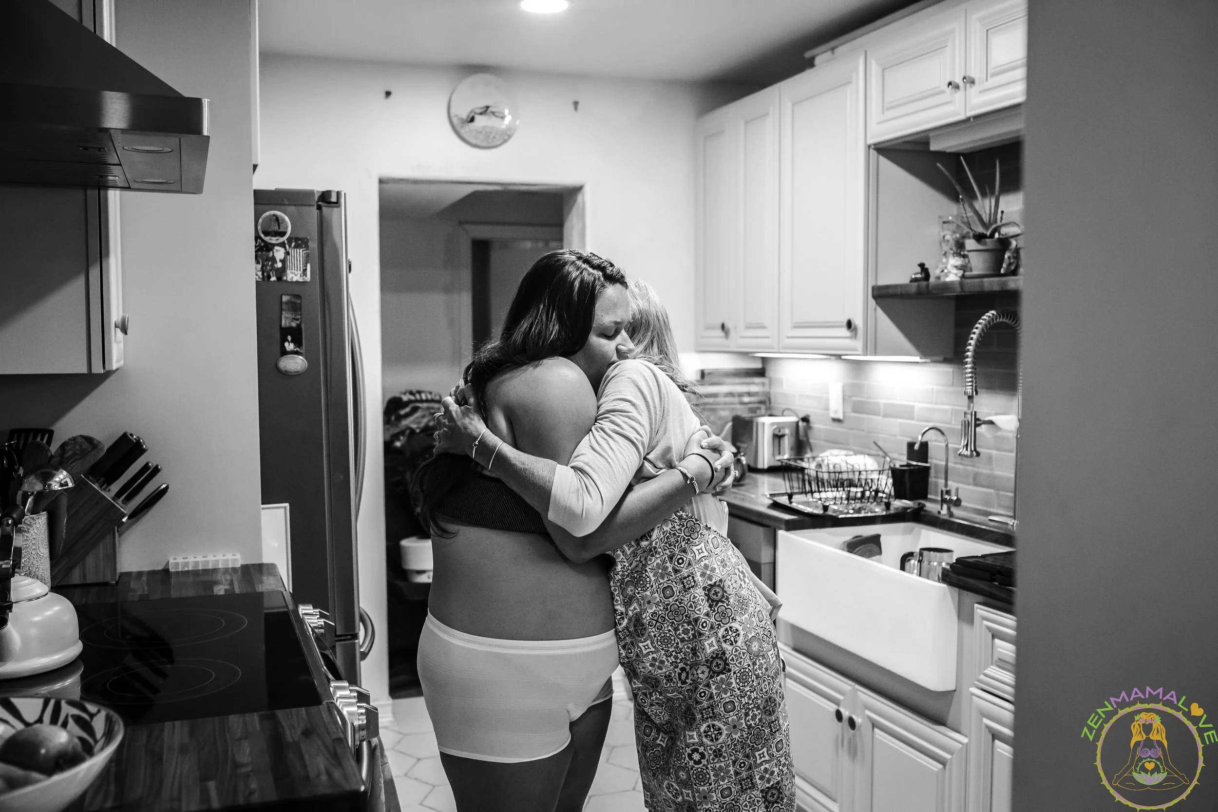 Two women hugging in a kitchen, one with long dark hair and the other with light hair, surrounded by kitchen cabinets and appliances.