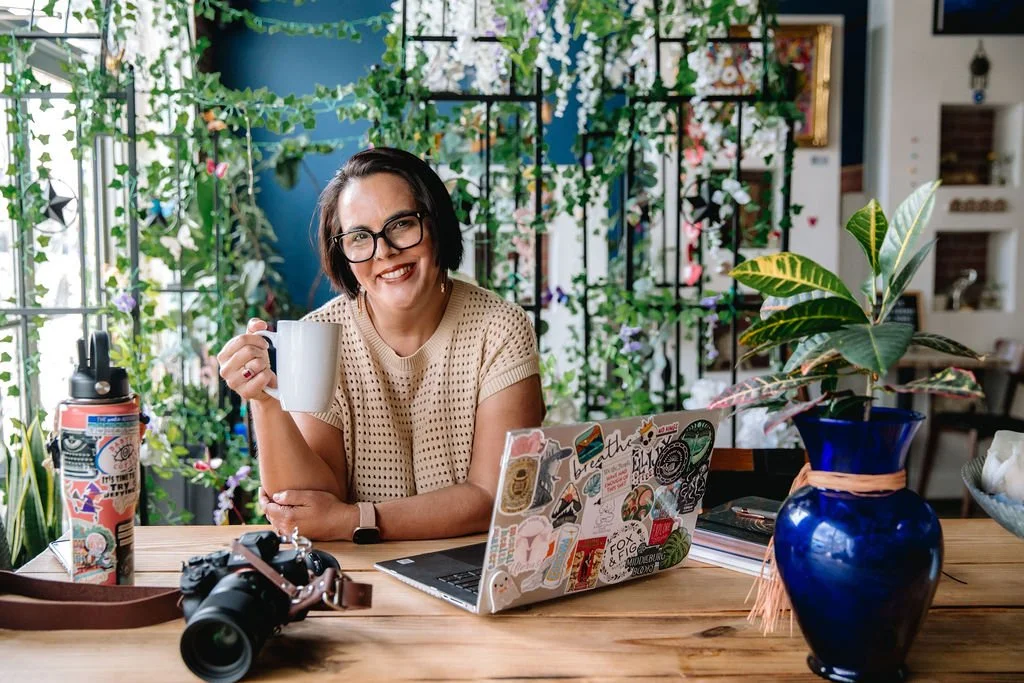 A smiling woman with glasses holding a white mug sits at a wooden table decorated with a laptop, a camera, a water bottle, and a blue vase with a green plant. The background features lush green plants and a window, creating a cozy indoor garden atmosphere.