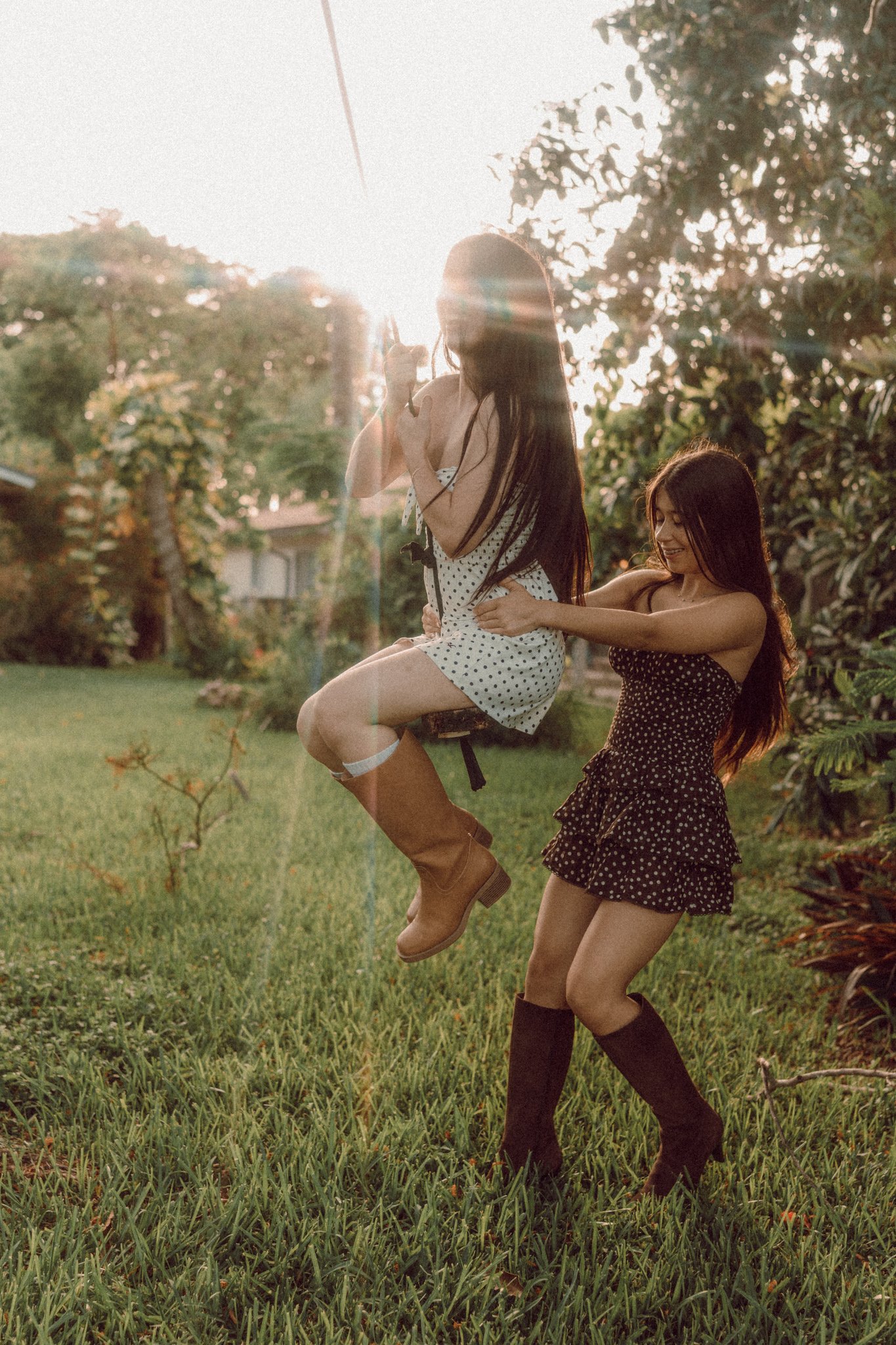 Two women playing on a swing in a garden during sunset, with lush greenery, trees, and a house in the background.