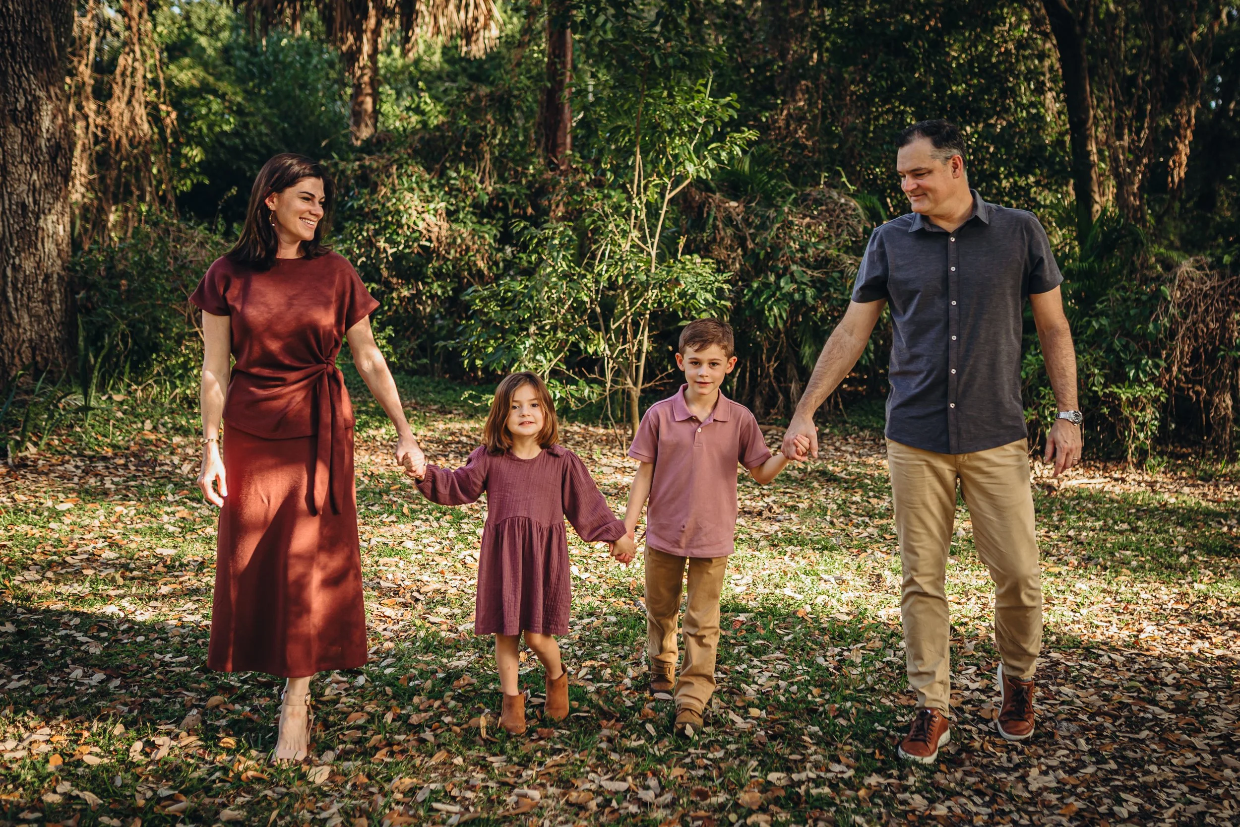 A family of five walking hand in hand through a wooded area with fallen leaves on the ground, smiling and enjoying a sunny day.