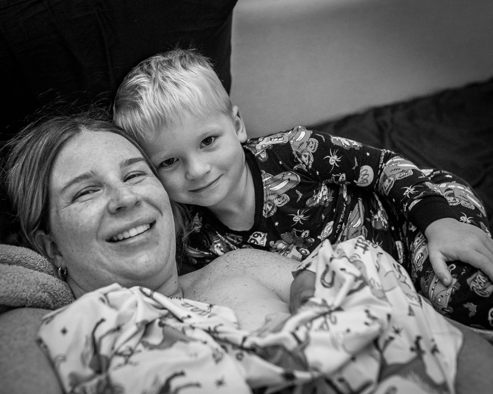 A young boy with blonde hair lies on a bed next to a smiling woman, likely his mother, with her face partially visible, both appearing happy and relaxed.