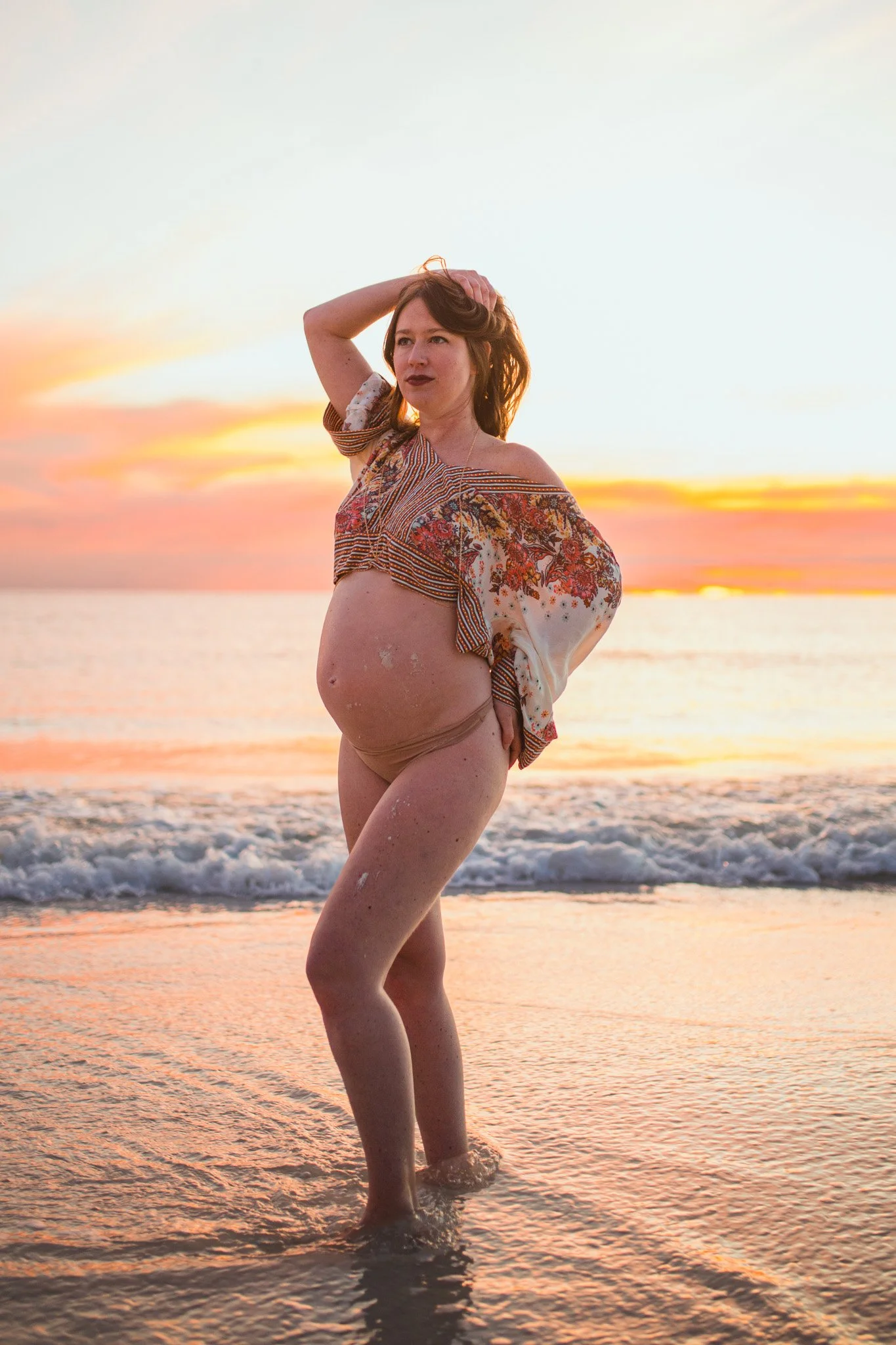 A woman standing in the ocean at sunset, with her hand on her head, wearing a patterned shirt and beige swim bottoms.