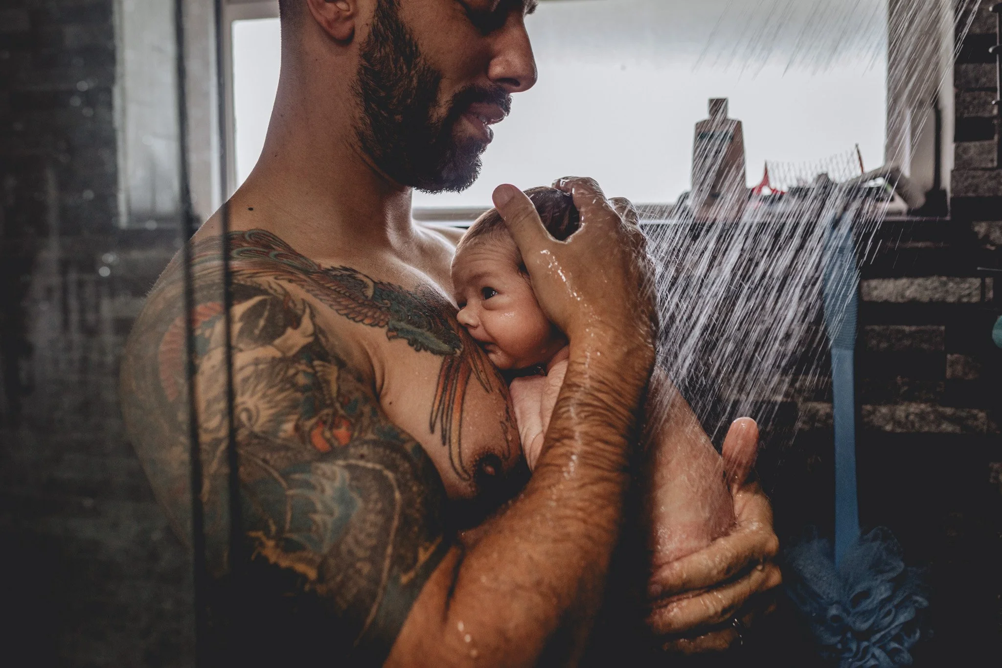 A man with tattoos on his arm and chest taking a shower, holding a baby close to him.