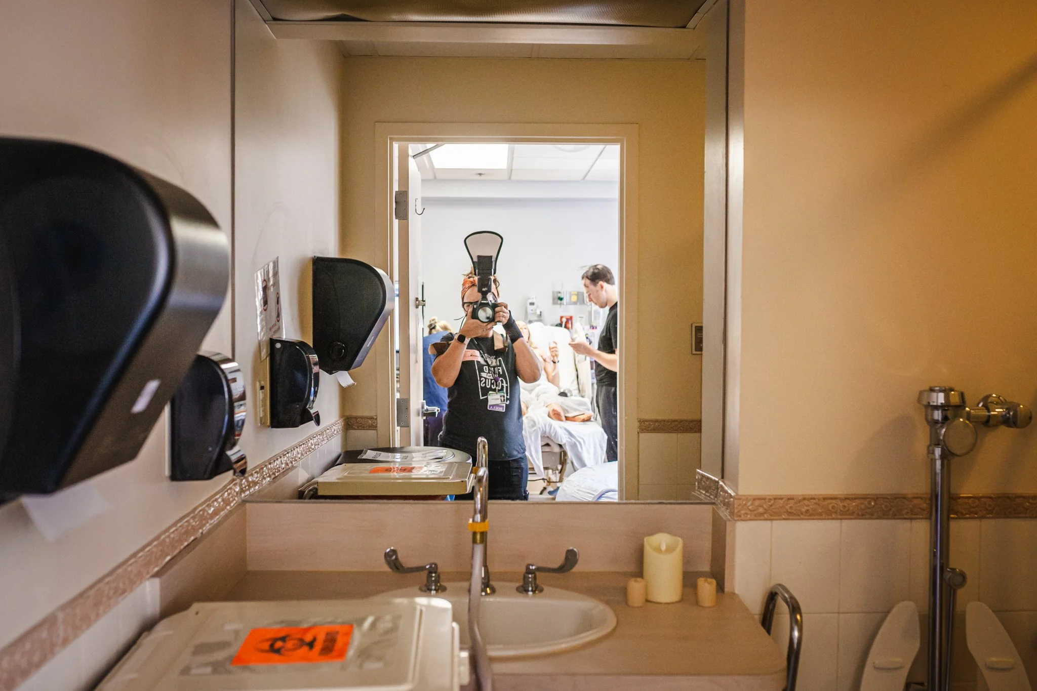 Hospital room with a person taking a photo of their reflection in a mirror, several patients in beds, medical equipment, and a person standing beside a bed, medical supplies on a countertop, and candles on the sink.