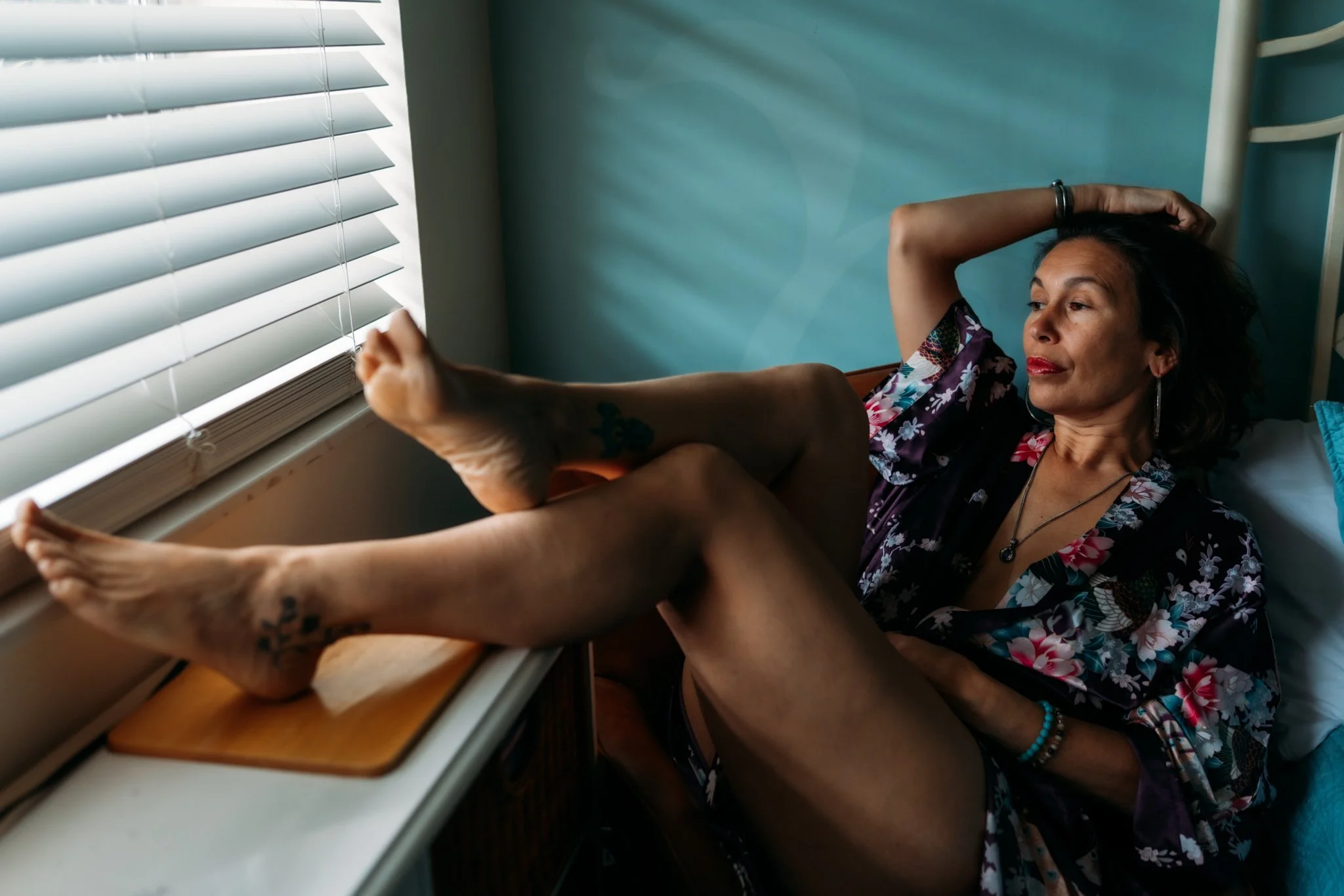 Woman with dark hair and tattoos, wearing a floral shirt, lying on a bed by a window with blinds, holding a smartphone.