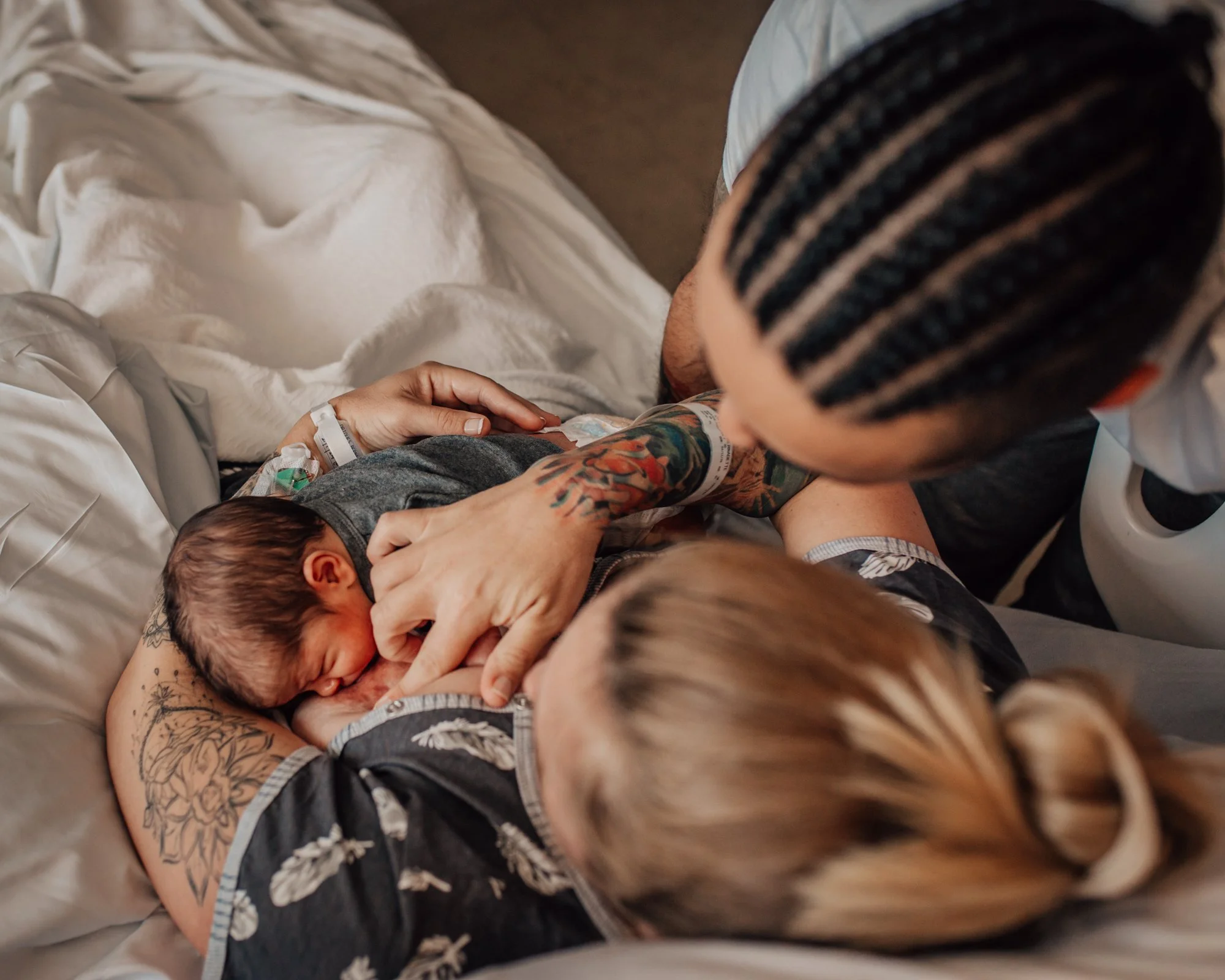 A person with tattoos and a striped beanie is lying on a hospital bed holding a newborn baby. The scene appears to be a birthing or postpartum moment in a hospital room.