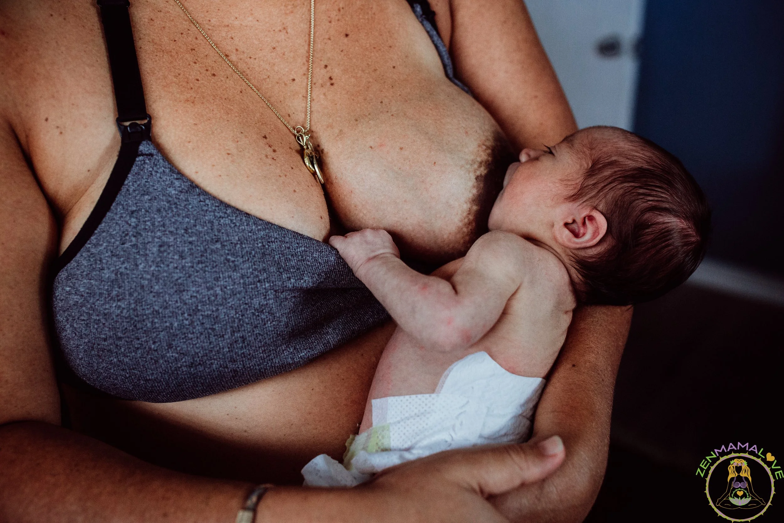 A woman breastfeeding a newborn baby with a diaper, wearing a blue top and a gold necklace, with a floral logo in the lower right corner.