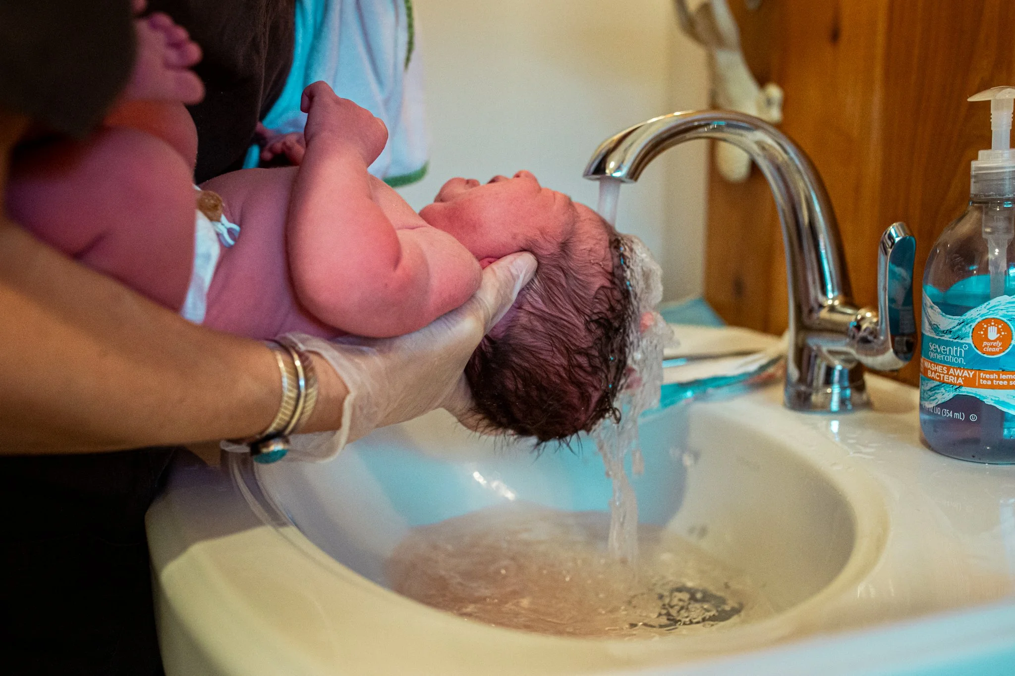 A newborn baby being washed in a sink by an adult, with soap and water running, and a bottle of baby wash nearby.
