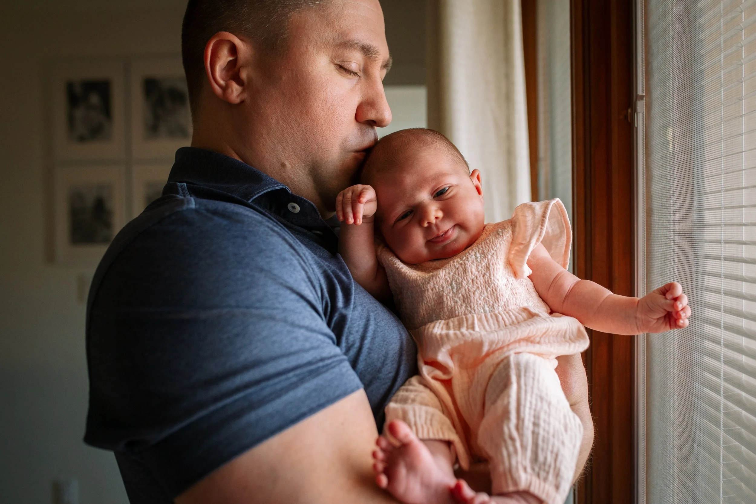 A man with short hair holding a smiling baby girl near a window with blinds, both looking outside.