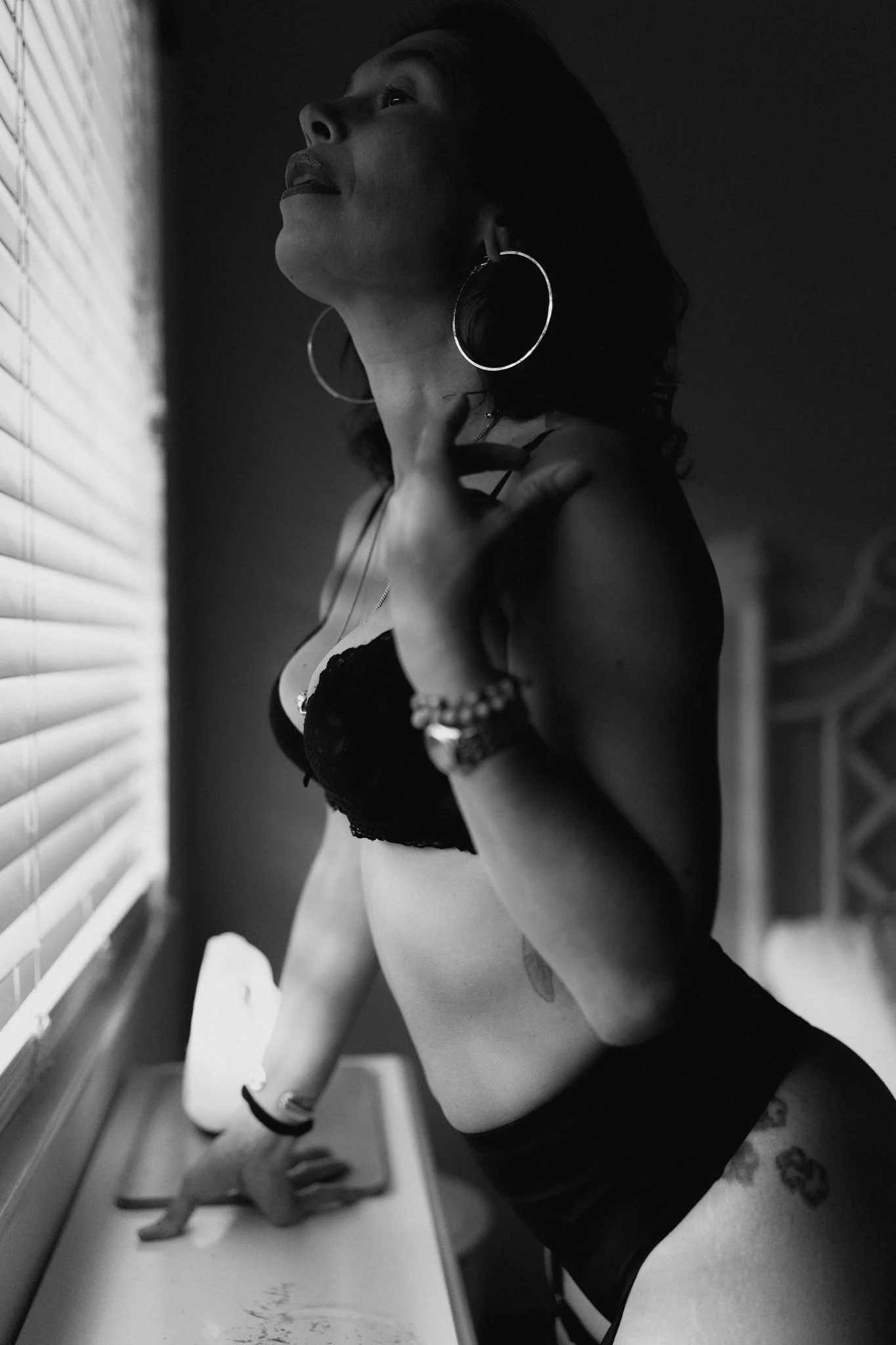 A woman with large hoop earrings and a necklace, wearing a black lace top and bracelets, leaning against a window with blinds, looking upwards in a contemplative pose.