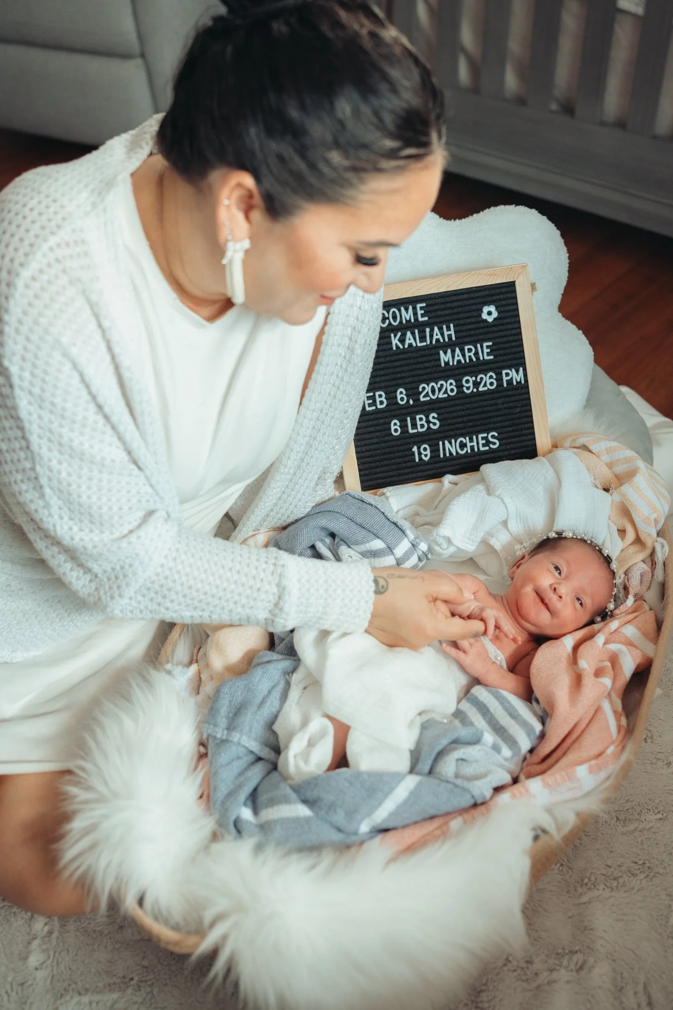 A woman with dark hair, earrings, and light-colored clothes is smiling and holding a baby wrapped in patterned clothes, laying on a soft blanket. There is a small black letter board behind them with the message welcoming Kaliah Marie, born February 6, 2026, at 9:26 pm, weighing 6 pounds and 19 inches long.