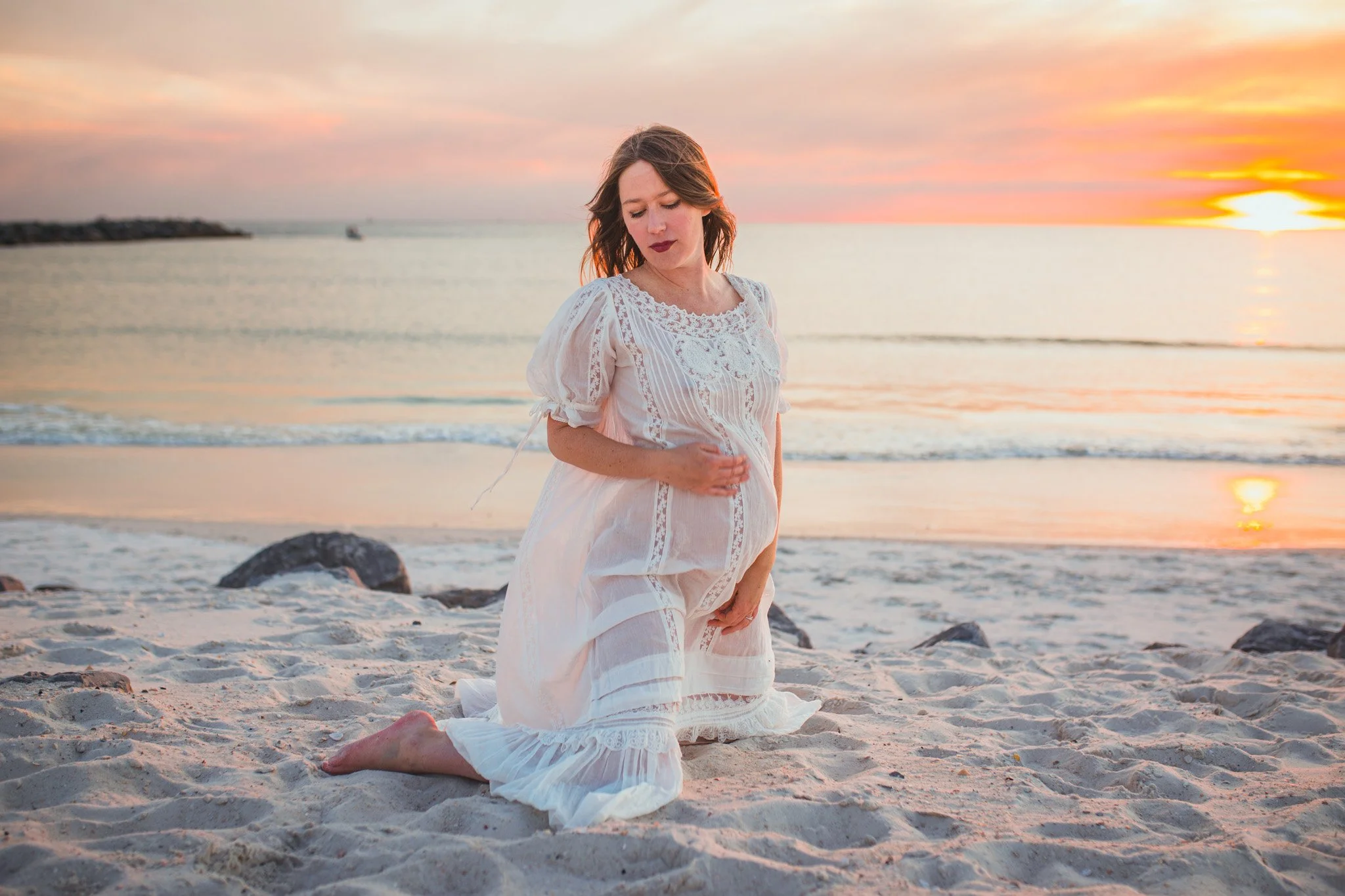 A woman kneeling on the beach during sunset, wearing a white, lace dress.