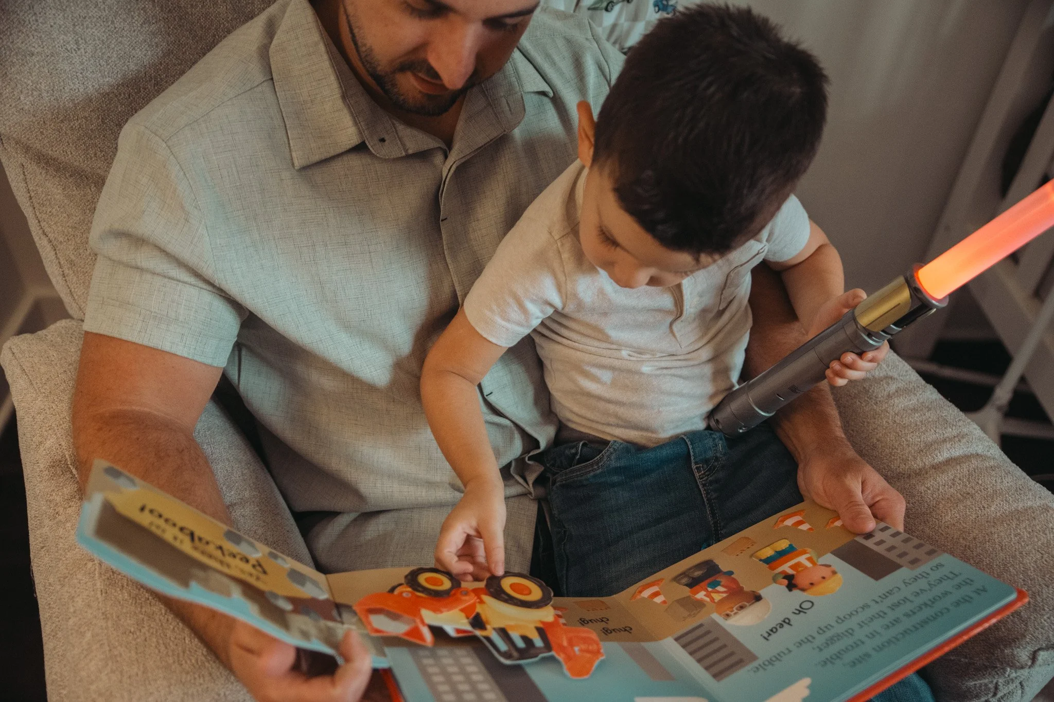 A man and a young boy sitting on a couch reading a book about cars, with the boy holding a toy car. The man is looking at the book while the boy looks at the toy car.