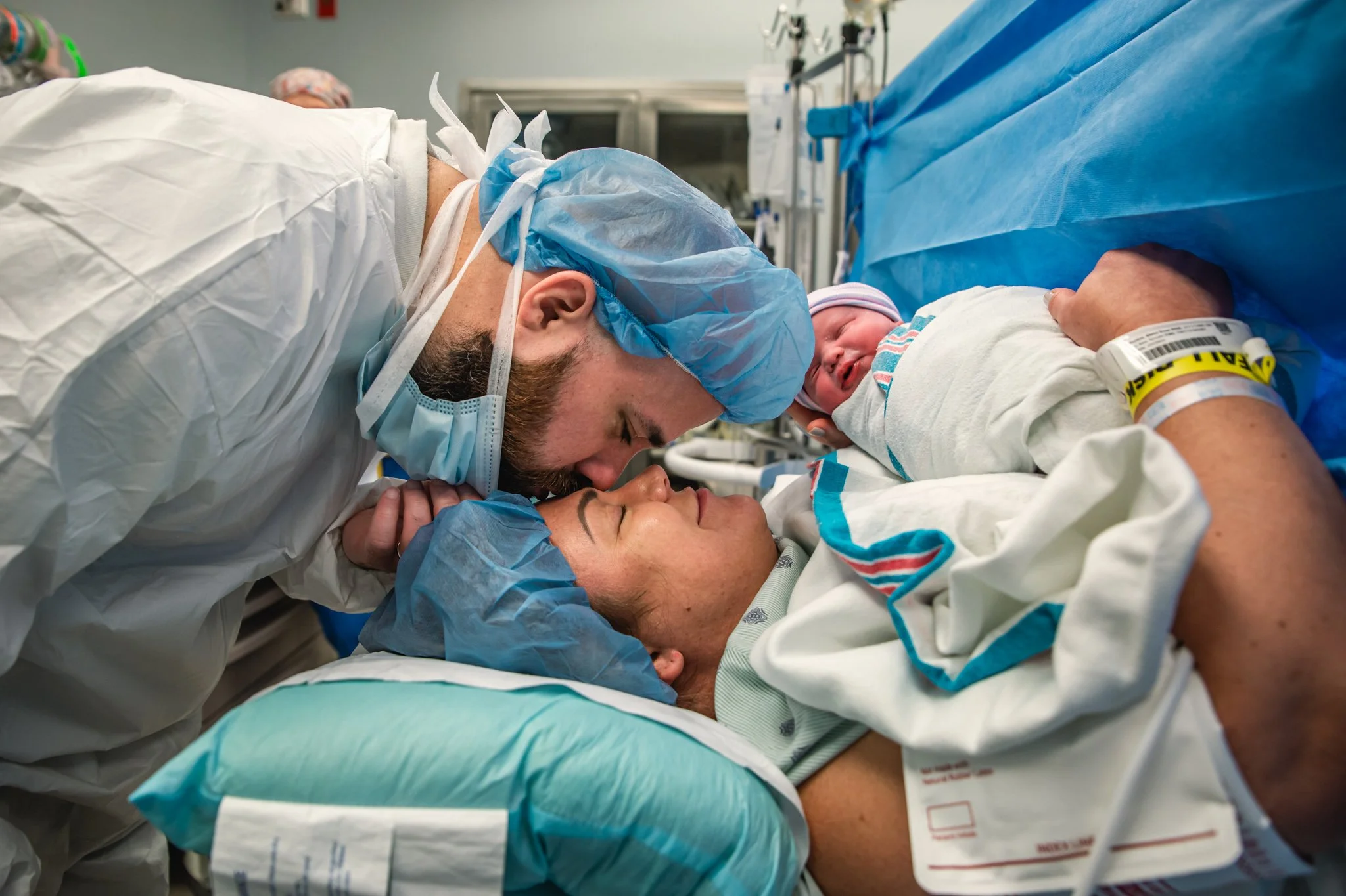 A man in a hospital bed holding and kissing a woman while a newborn baby sleeps between them, all dressed in hospital attire.