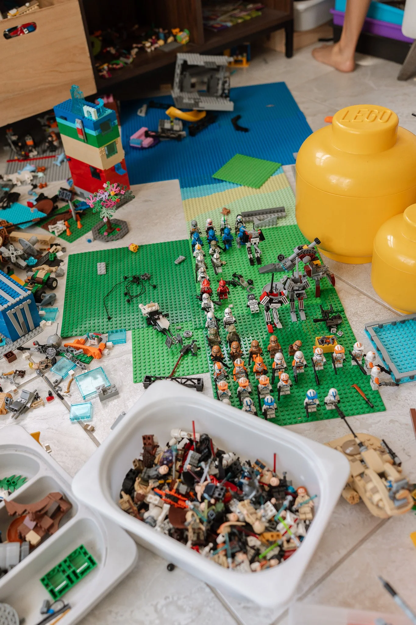 A messy scene of LEGO bricks, minifigures, and assembled LEGO structures on a tiled floor, with large yellow LEGO storage containers and some partially completed LEGO projects visible.