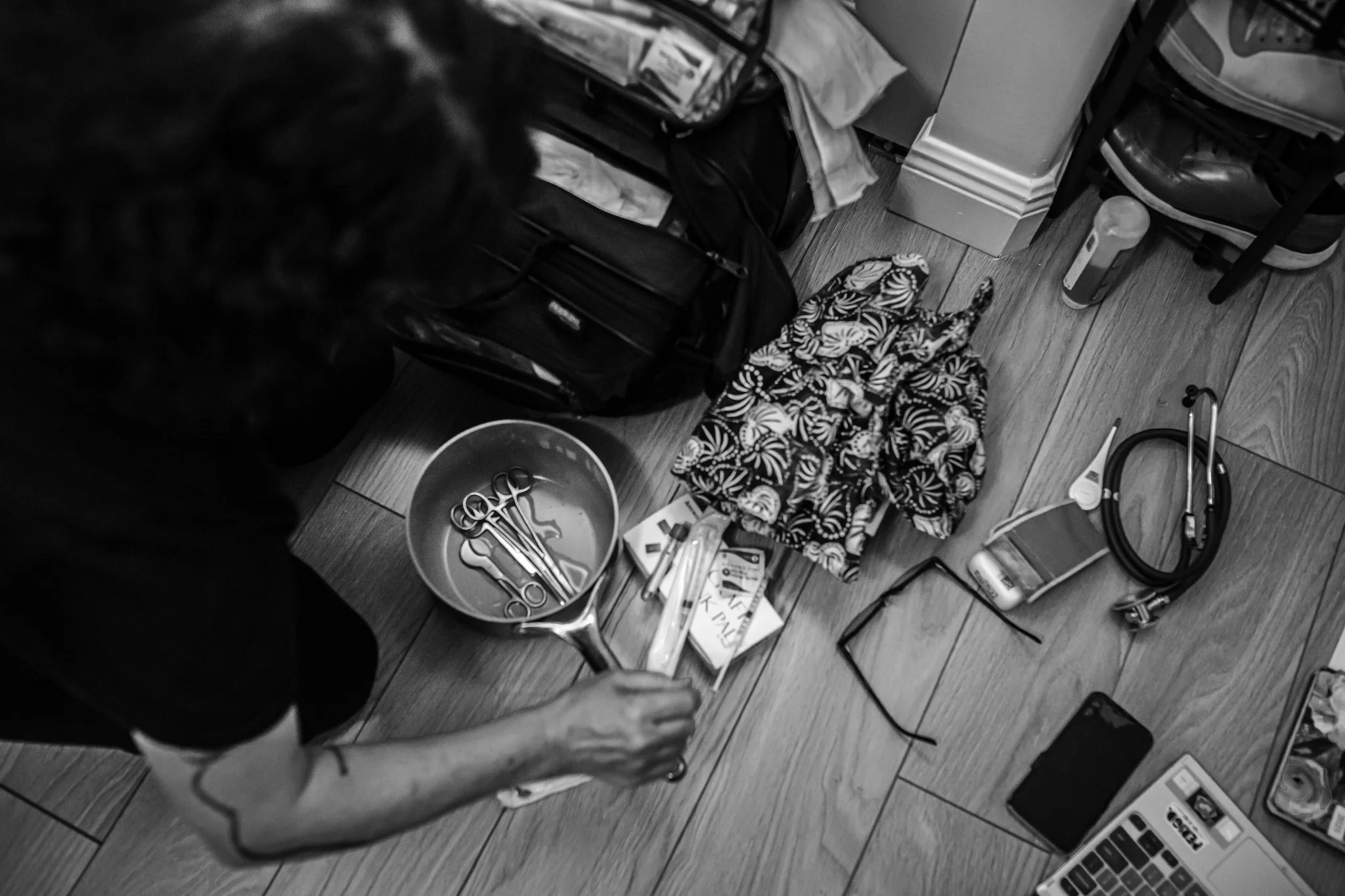 A cluttered workspace with a person holding a pair of scissors, surrounded by various objects including a bowl with more scissors, a patterned cloth, a laptop, skincare or cosmetic products, and several containers, on a wooden floor.