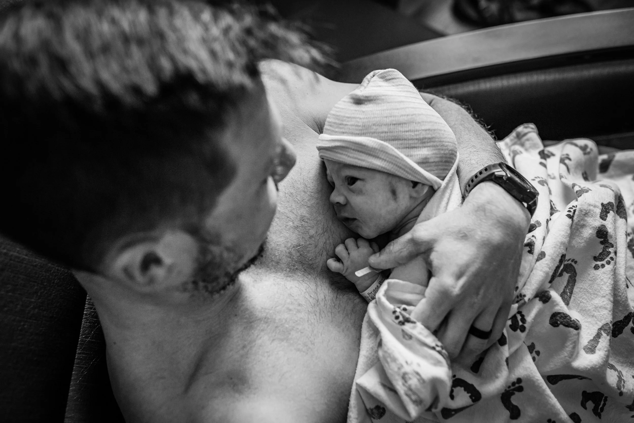 A newborn baby wearing a hat and patterned blanket, being held close to a woman's chest, while a man looks on. The scene appears to be in a hospital or a home setting, with the woman and the man showing tender interest towards the baby.