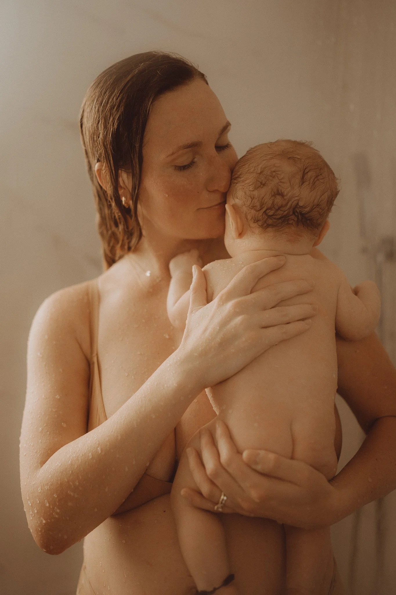 A woman with wet hair and water droplets on her skin holding a naked baby with curly hair, both in a warm-toned setting.
