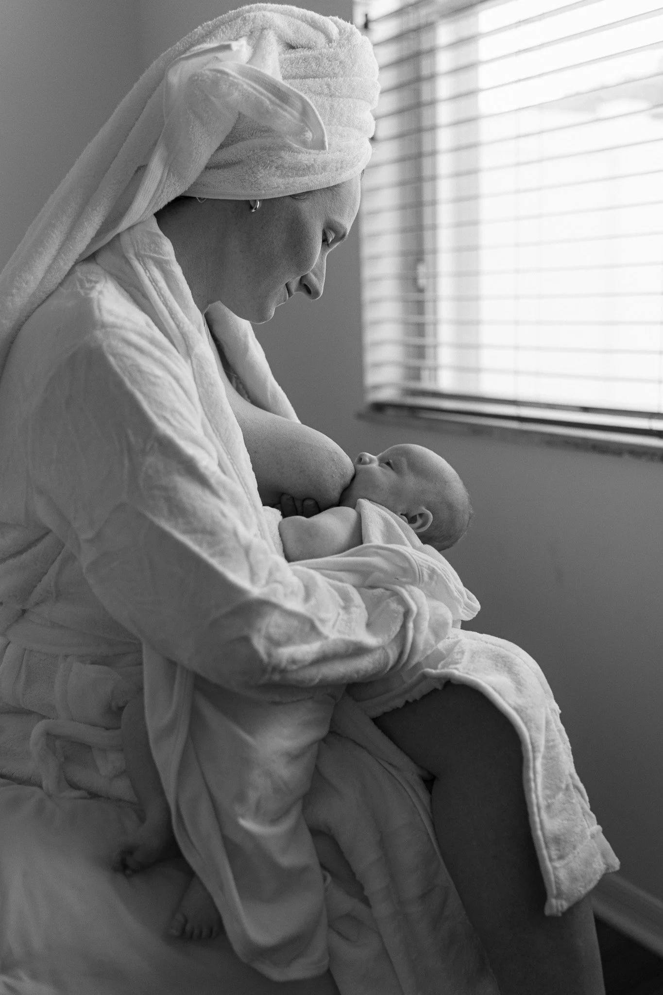 A woman with a towel wrapped around her head, wearing earrings, holding a baby and breastfeeding, near a window with blinds.
