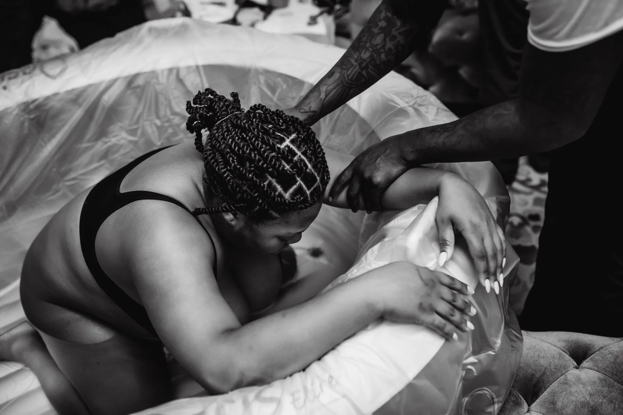 A woman with braided hair and tattoos lying on a bed, with a person assisting or touching her arm. The photo is in black and white.