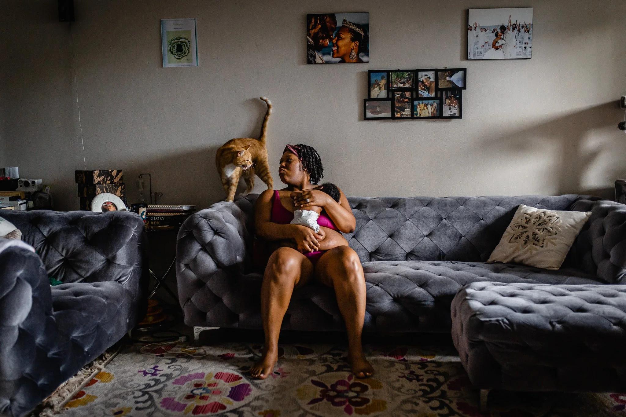A woman sitting on a gray tufted sofa in a living room, holding a baby in her lap. There is an orange tabby cat on the back of the sofa next to her, with its tail raised. The room has framed photos and artwork on the wall, books, and various items on